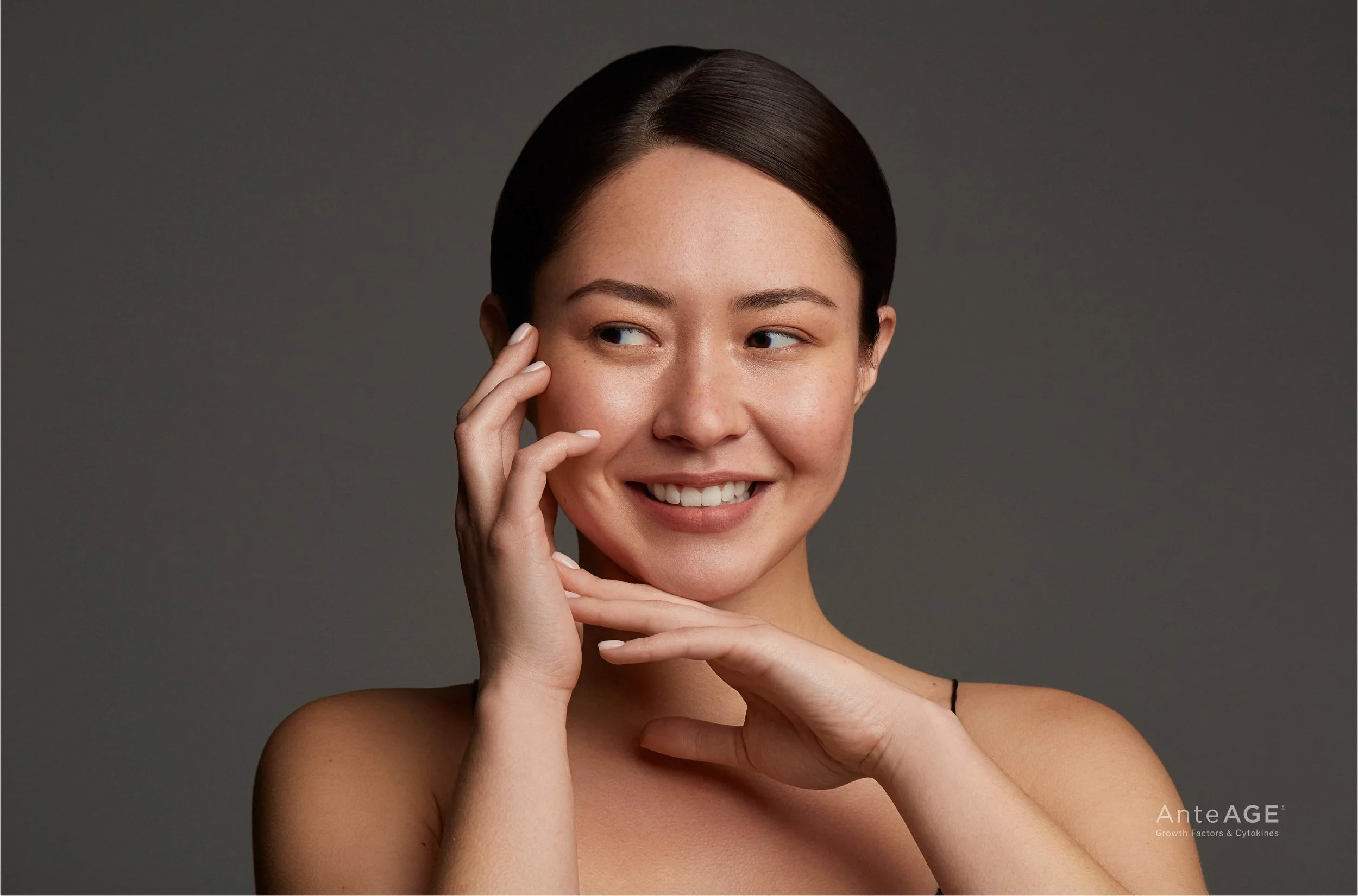 A young woman with smooth, clear skin smiling lightly, touching her face and chin, against a gray background.