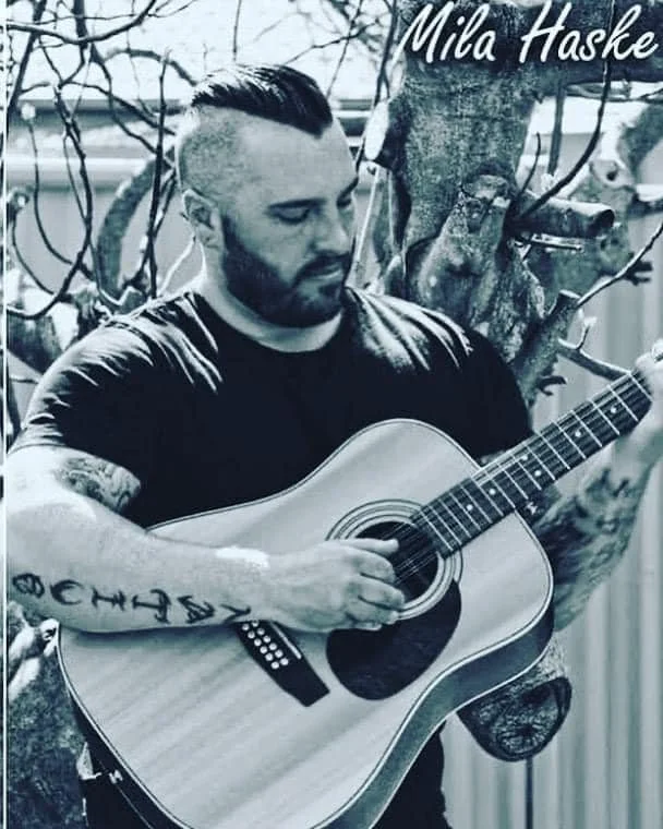 A man with a tattoo sleeve playing an acoustic guitar outdoors next to a tree.