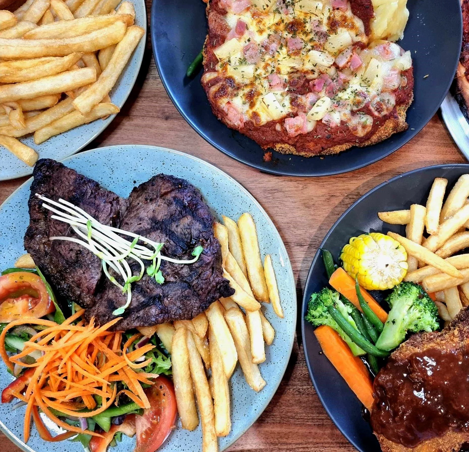 A meal featuring a grilled steak with fries and a side salad, a slice of pizza with ham and pineapple, and a plate with fried fish fillet topped with brown gravy, vegetables, and fries.