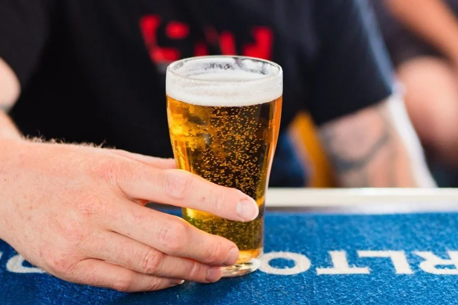 A person with a light skin tone holding a pint glass of golden beer with bubbles and a foamy head on a bar counter.