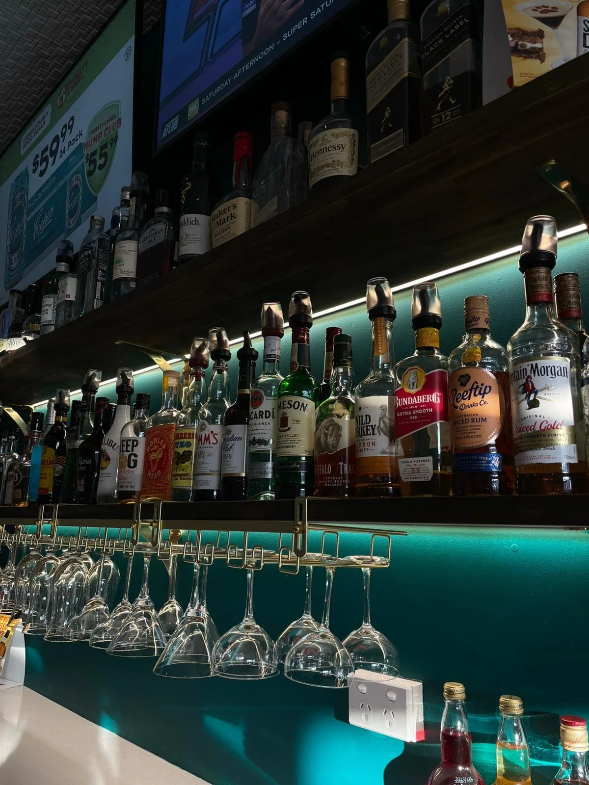 Bar shelf with various bottles of liquor and upside-down wine glasses hanging below, illuminated by teal backlight with a TV screen above.