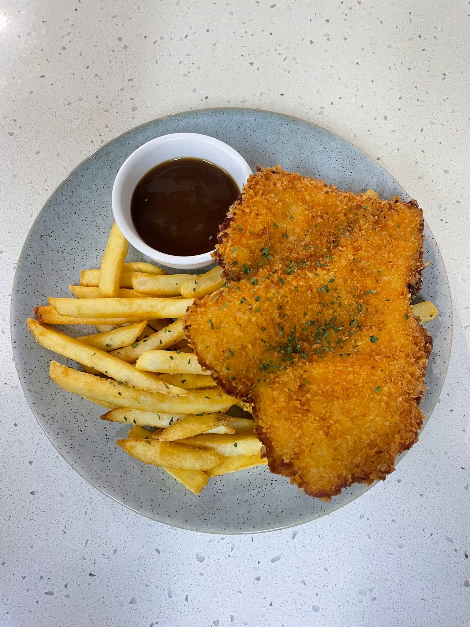 Plate with breaded fried fish fillets, French fries, and a small cup of dark dipping sauce on a light-colored speckled surface.