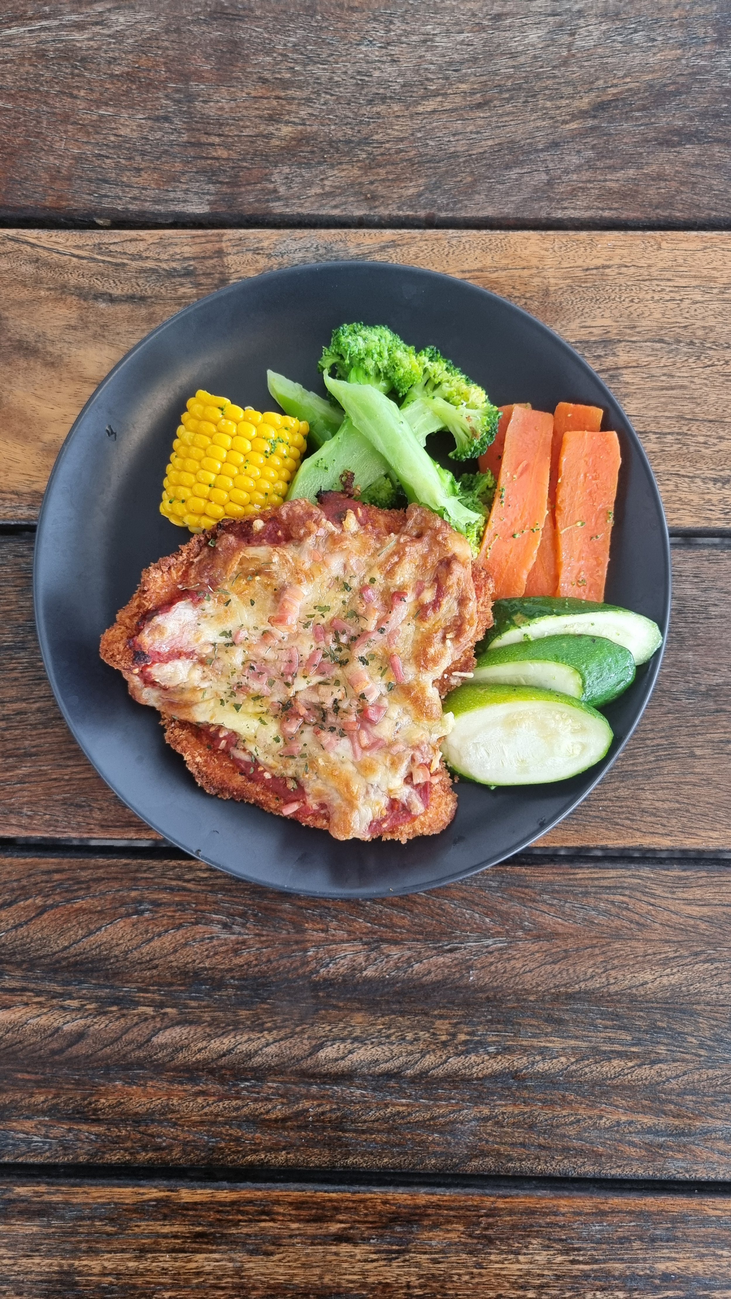 Plate with fried chicken topped with cheese and bacon, accompanied by corn on the cob, broccoli, carrots, and cucumber slices on a wooden table.