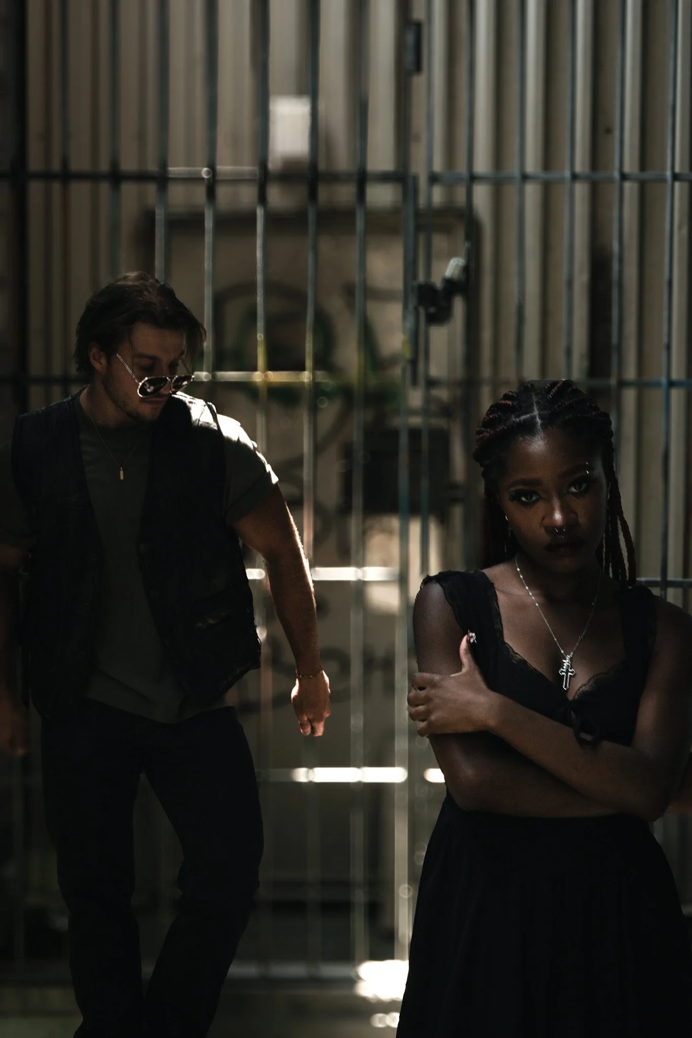 A man and woman standing close together in an industrial setting. The man, head down, looking at girl, with dark hair and a beard, has a serious expression and wears a black leather vest. The woman, with braided hair and dark makeup, has a serious ex