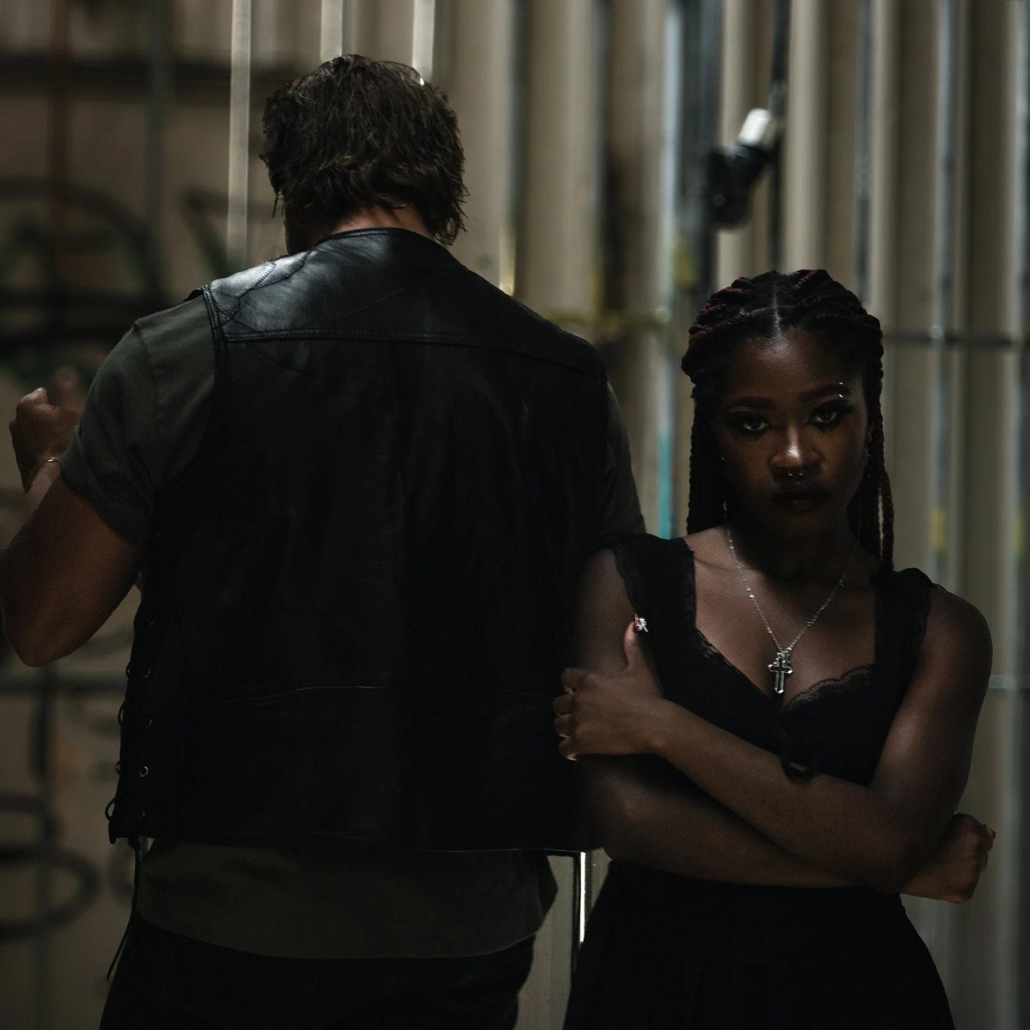 A man and woman standing close together in an industrial setting. The man, back to the scene, turned to the bars, with dark hair and a beard and wears a black leather vest. The woman, with braided hair and dark makeup, has a serious expression and we
