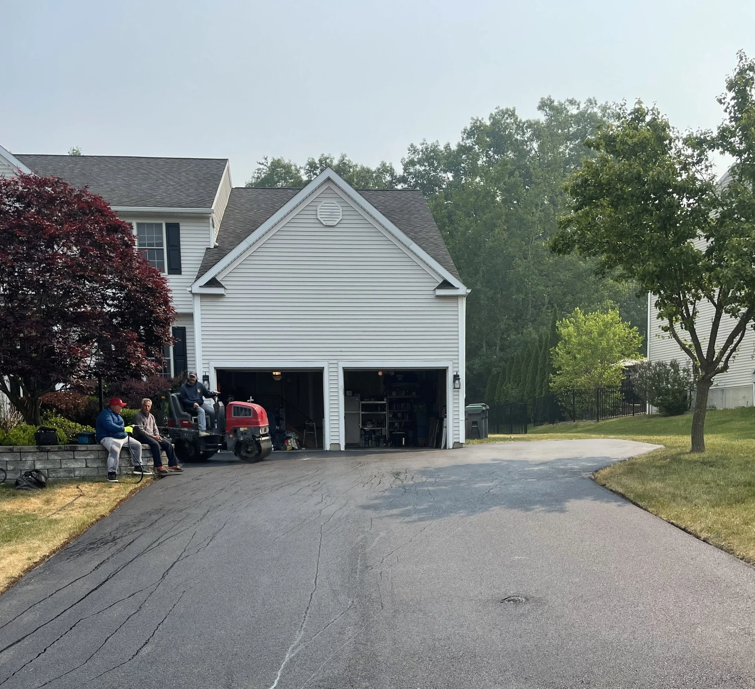 A suburban house with a two-car garage and an open driveway. Two people are sitting on a garden wall beside a tree, and another person is driving a riding lawn mower. The garage doors are open, revealing storage shelves inside. It's a cloudy day.