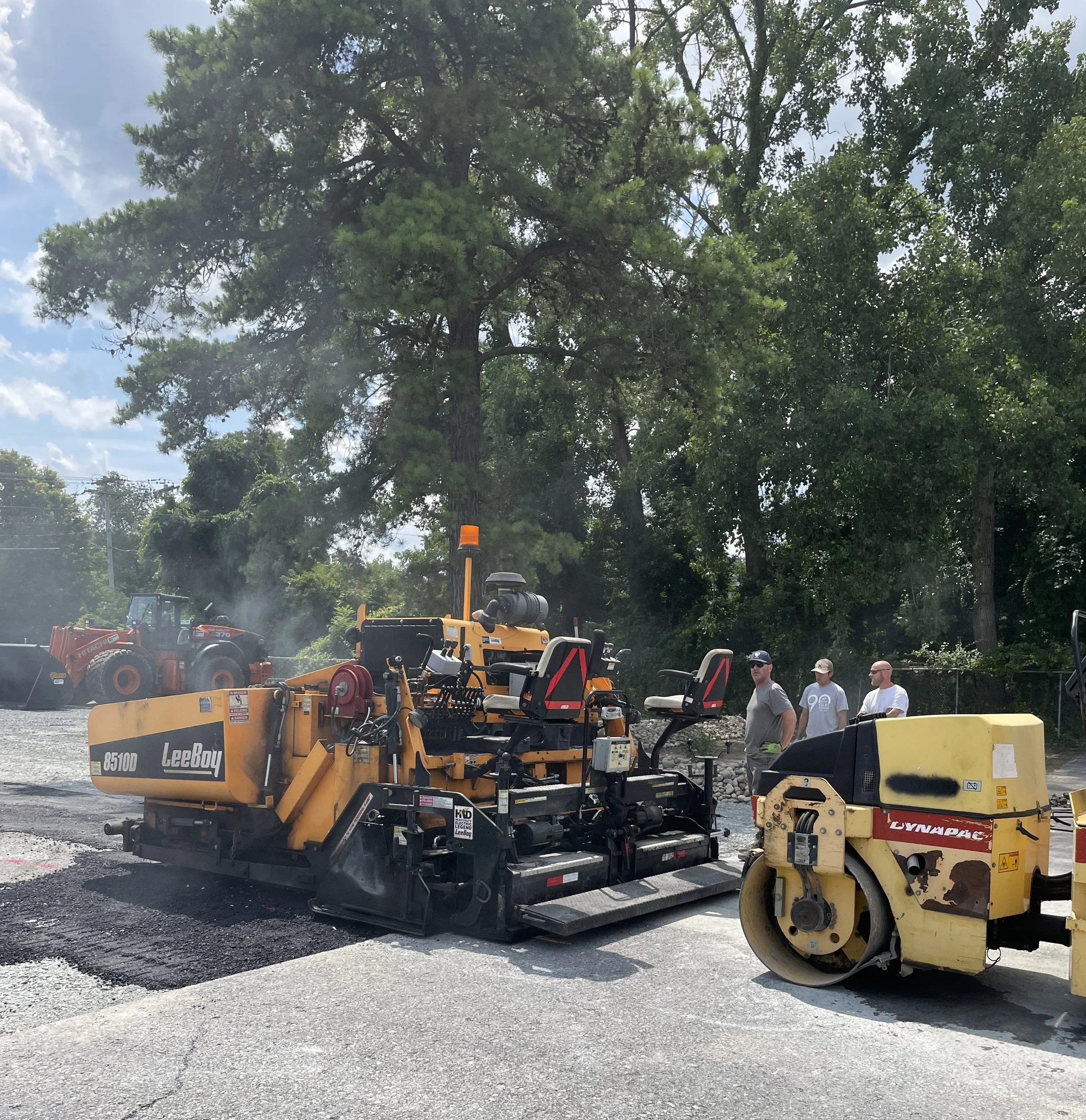 Paving machinery and workers on a construction site with trees in the background.