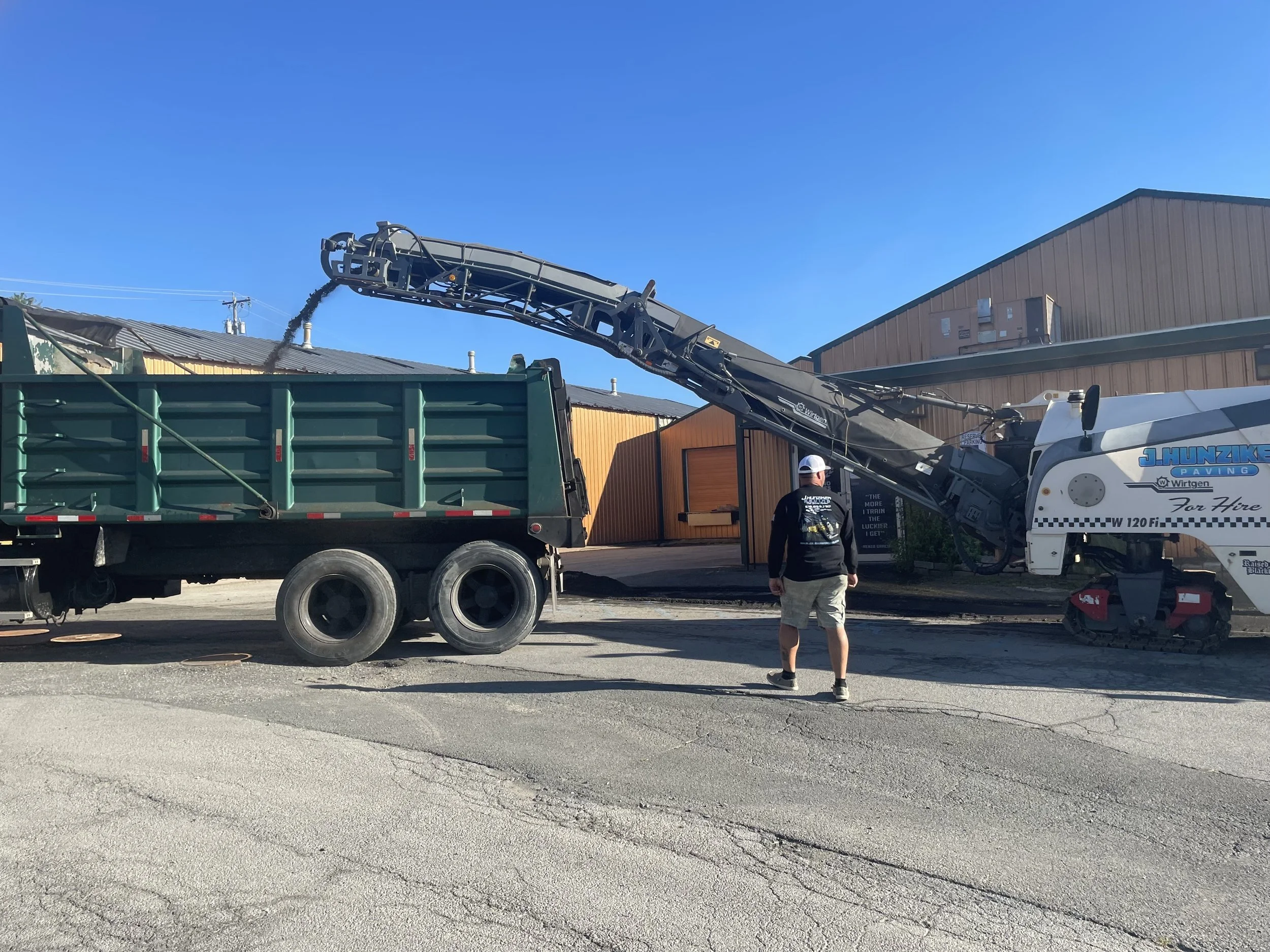 Construction site with an asphalt milling machine unloading material into a dump truck, a man standing nearby, and industrial buildings in the background on a sunny day.