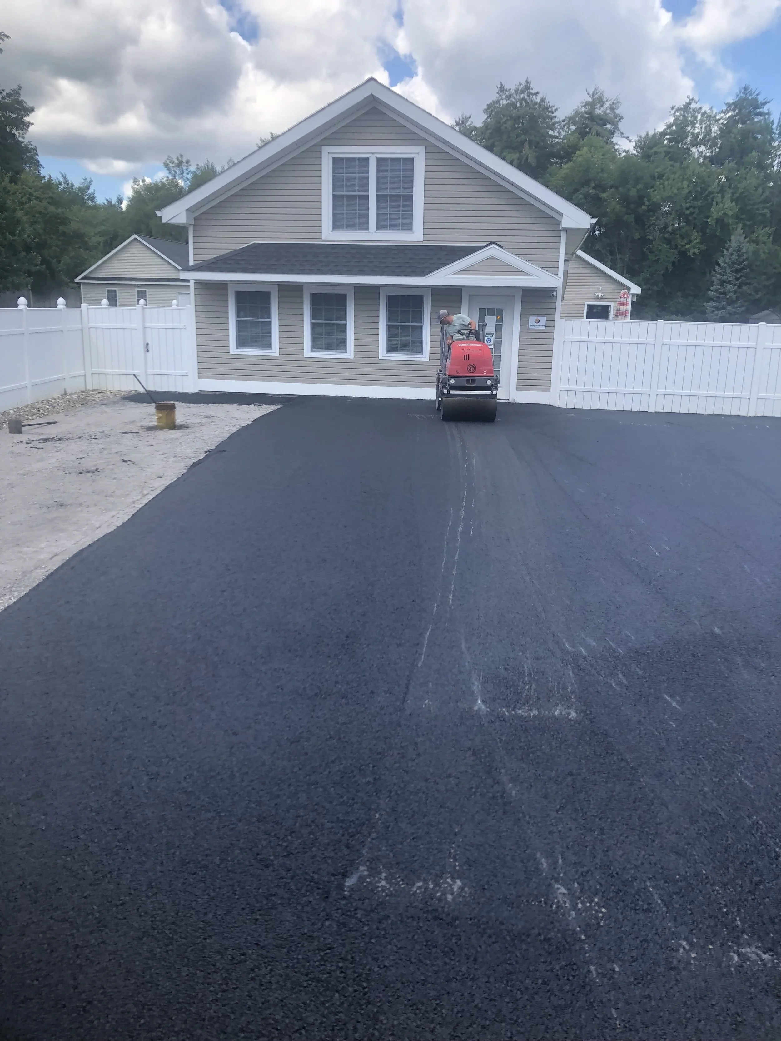 Newly paved driveway in front of a beige house with white trim and a red steamroller machine on the asphalt, surrounded by a white fence and trees in the background.