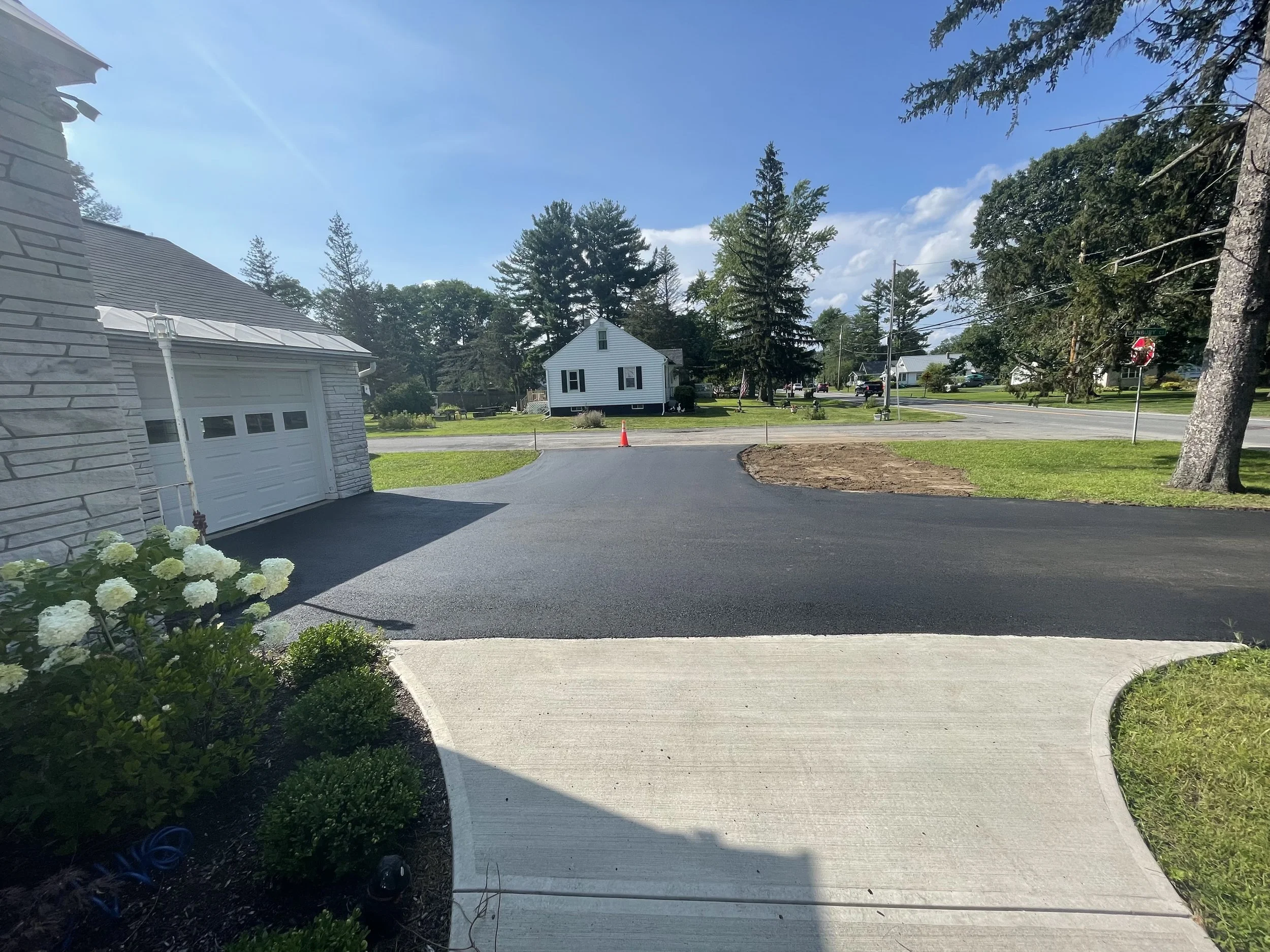 Suburban driveway with new asphalt, adjacent to a house with a garage, trees lining the street, and a clear blue sky.