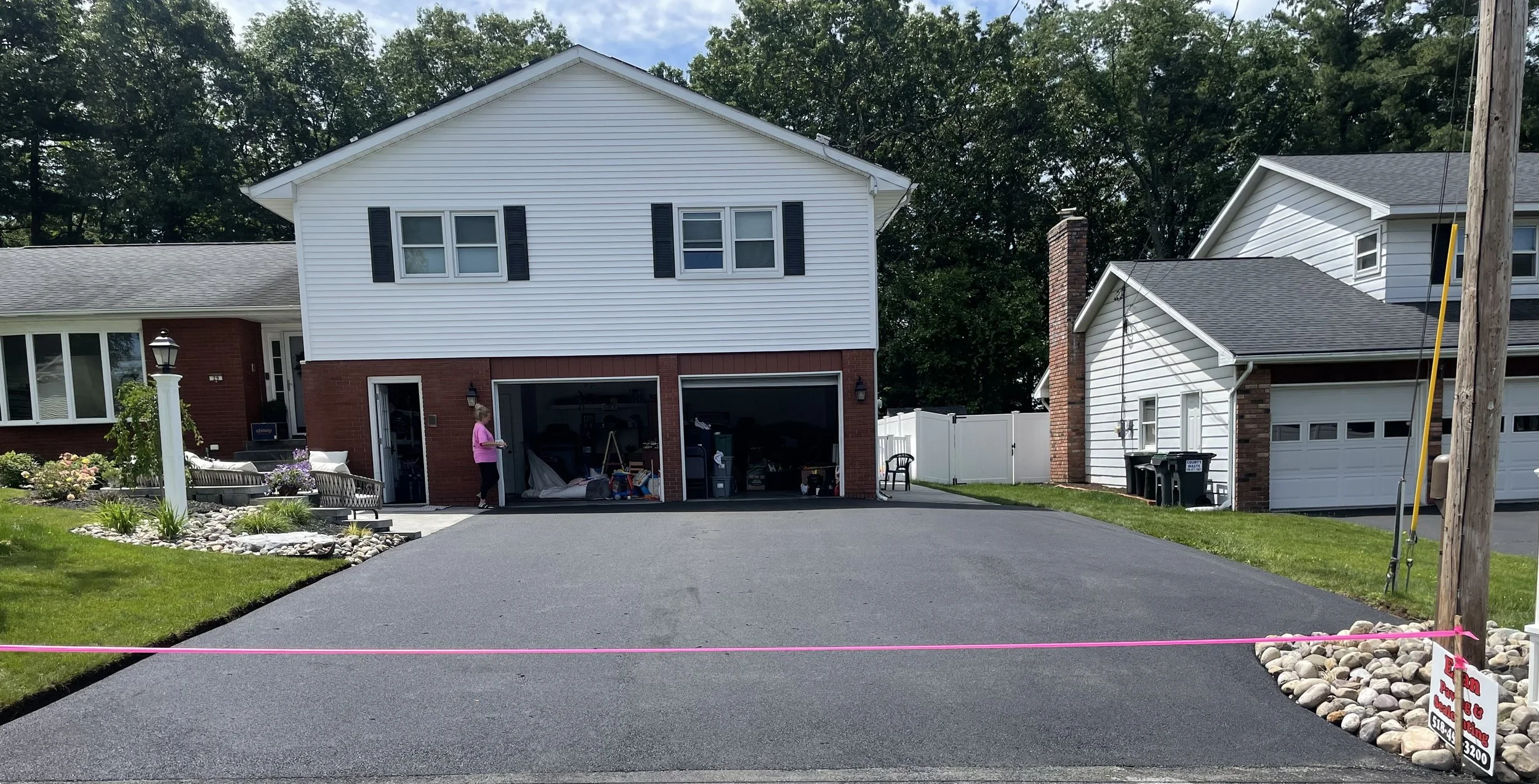 Two-story suburban house with open garage, paved driveway, and surrounding greenery. Pink caution tape blocks driveway access. A person stands near the entrance.