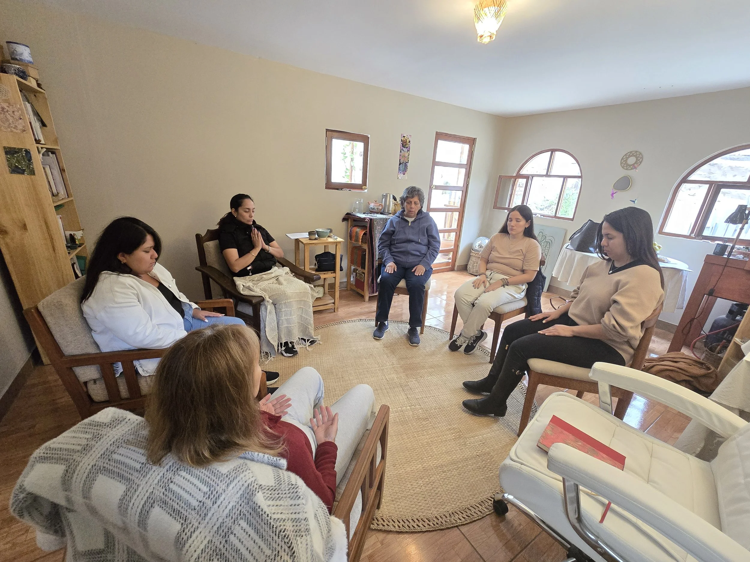 A group of women sitting in a circle in a cozy room, participating in a Reiki training, with some women having their eyes closed and hands in prayer.