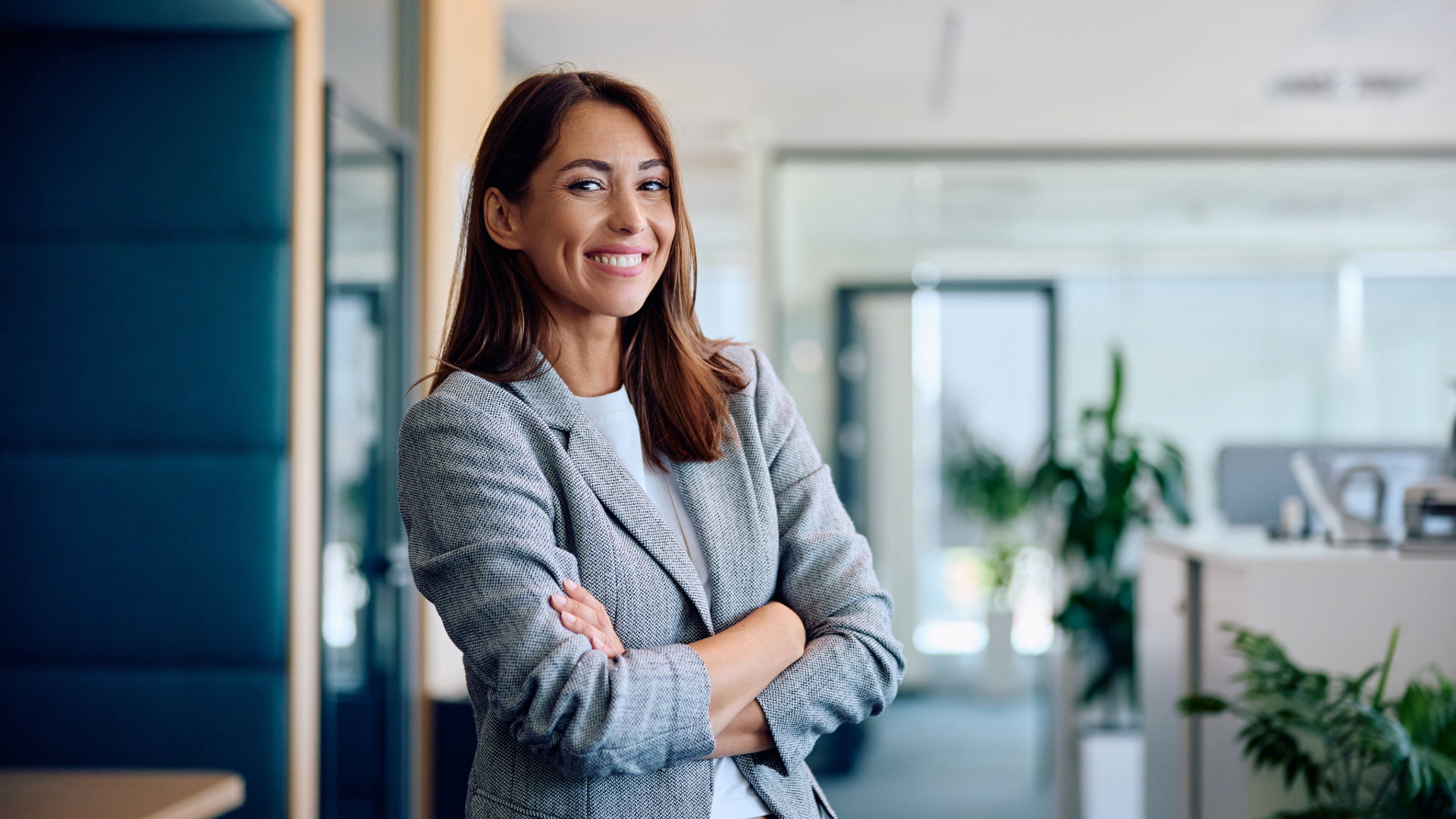 A woman with brown hair, smiling, wearing a gray blazer, standing with arms crossed in a modern office environment with plants and desks in the background.