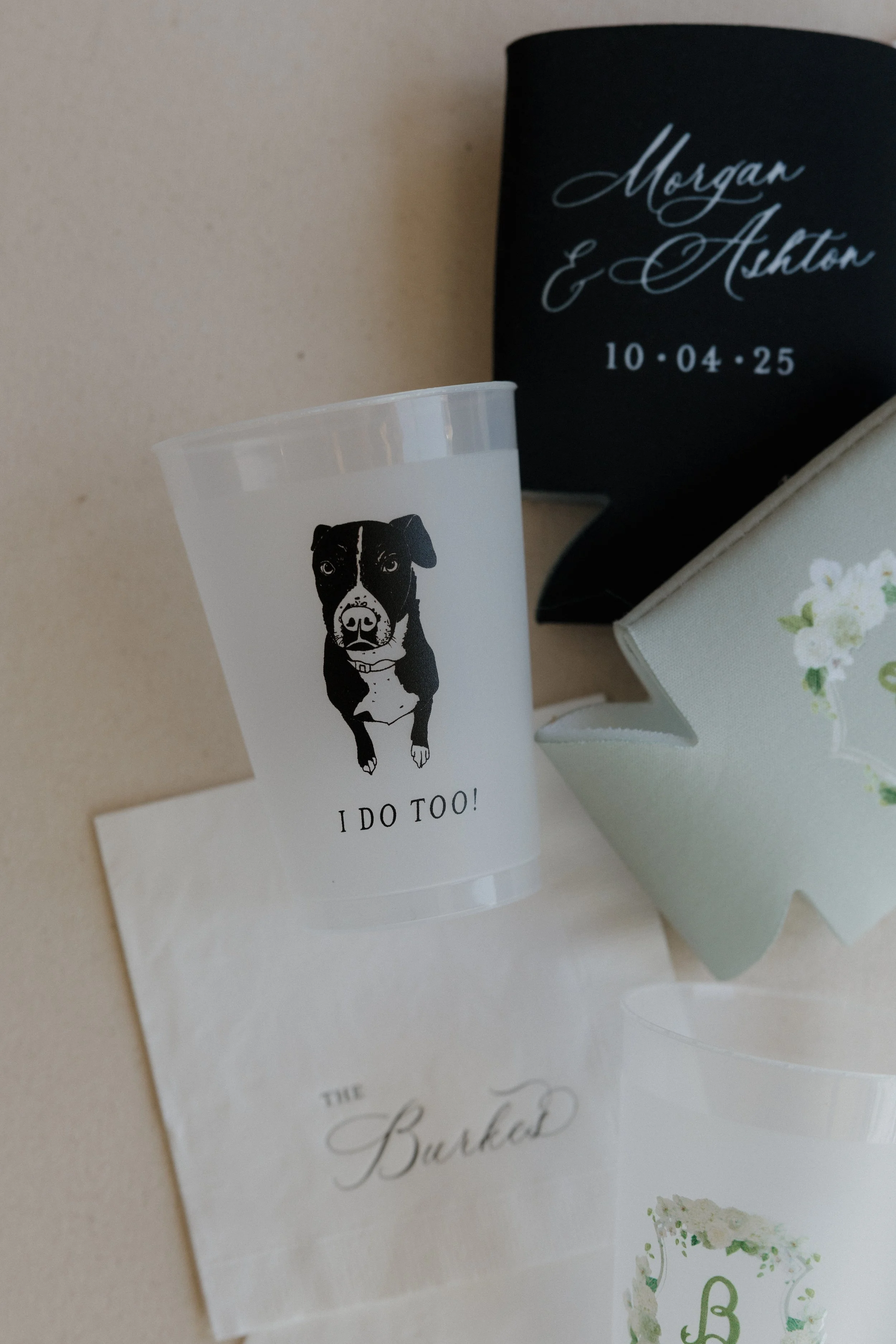 A plastic cup with a dog illustration and the text "I DO TOO!" on a table or surface, alongside a napkin, a white box with a floral design, and a black card with white cursive writing and a date.