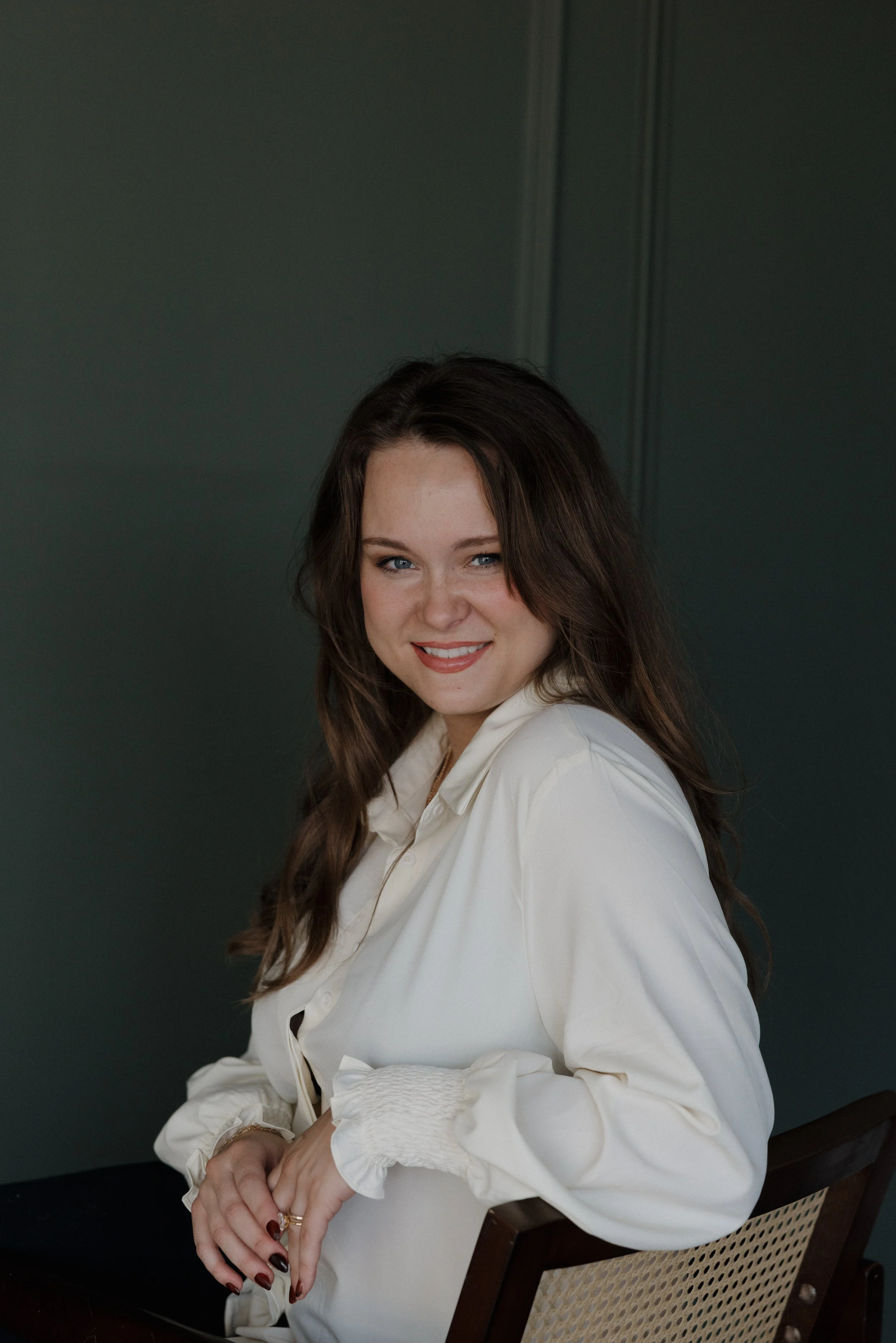 A woman with long brown hair, wearing a white blouse, smiling and sitting on a chair against a dark green wall.