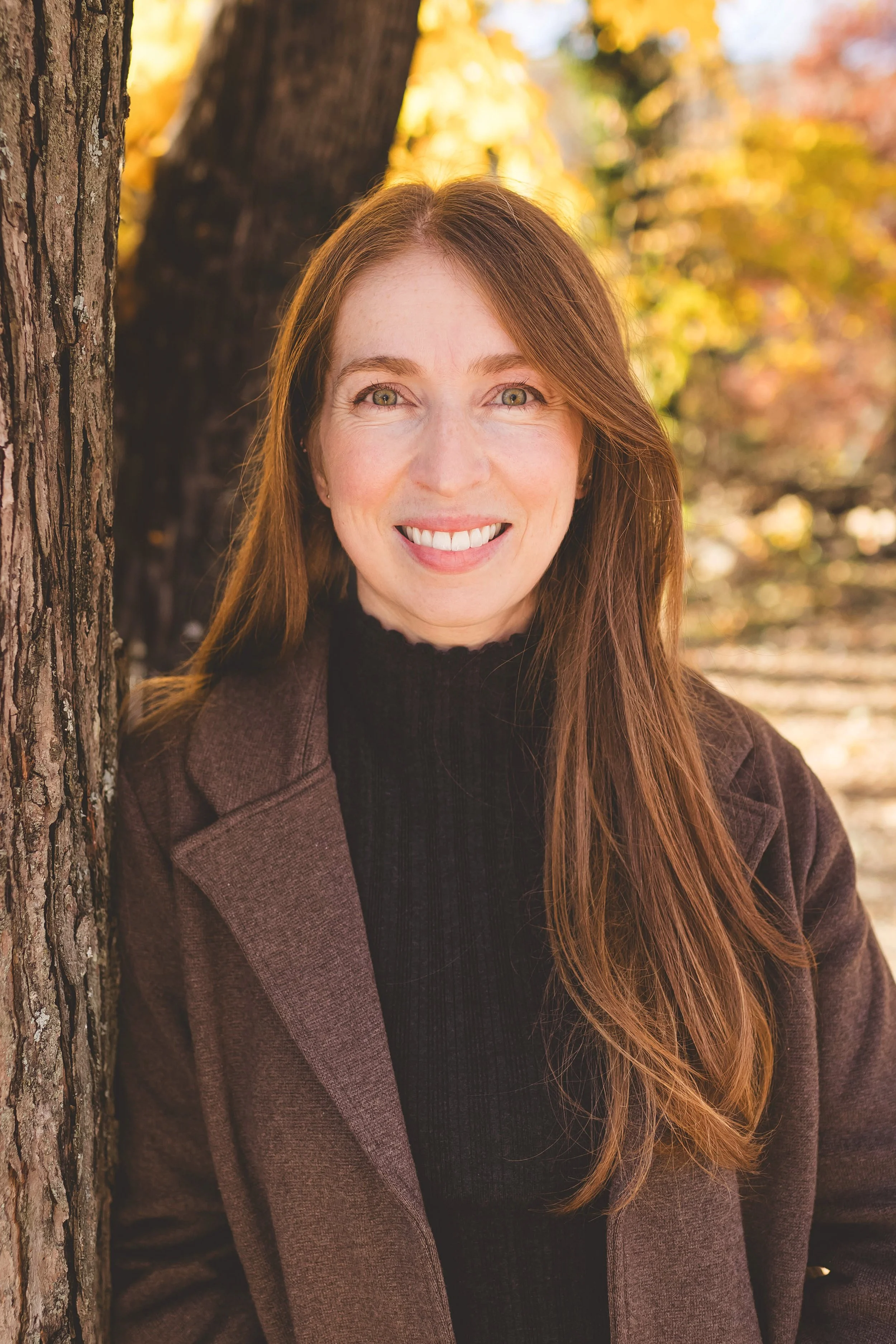A woman with long red hair, wearing a dark turtleneck and a brown coat, smiling outdoors in autumn with colorful fall foliage in the background.