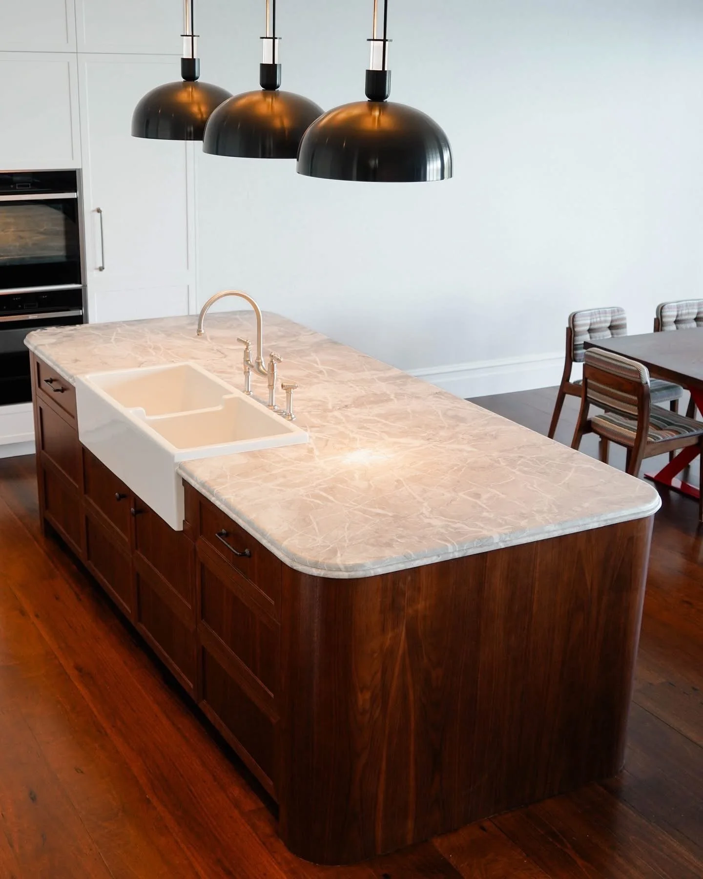 A modern kitchen island with a marble countertop, a double-basin white sink, and a bronze faucet. Overhead, three black pendant lights hang in a row. To the right, there are dining chairs with patterned upholstery around a table. The kitchen has whit
