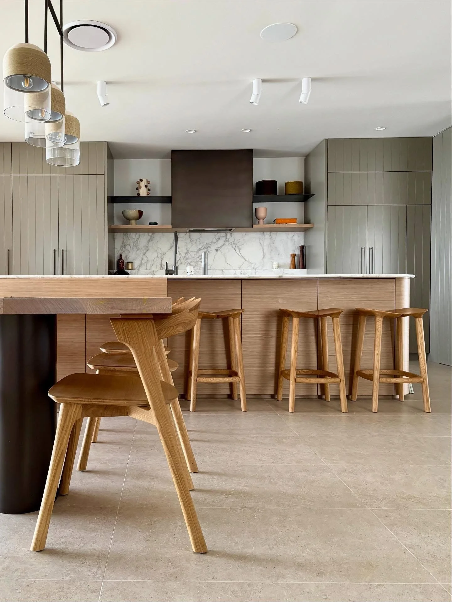 Modern kitchen with beige cabinetry, marble backsplash, wooden bar stools, and a wooden table with matching chairs. Overhead lighting features multiple pendant lights with beige and transparent shades.