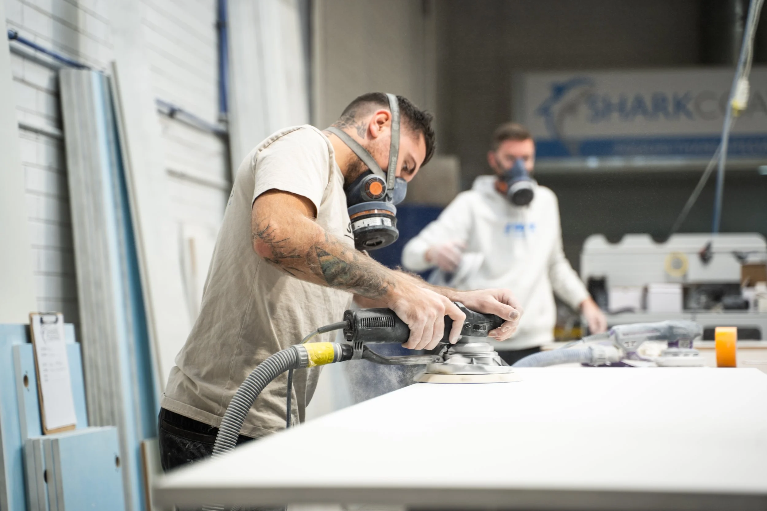 A man with tattoos and a mask working on a project with power tools in a workshop, with another person in the background.