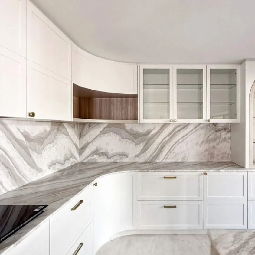 White kitchen cabinets with gold handles, marble backsplash with gray and beige patterns, and a marble countertop, with open glass cabinet doors.