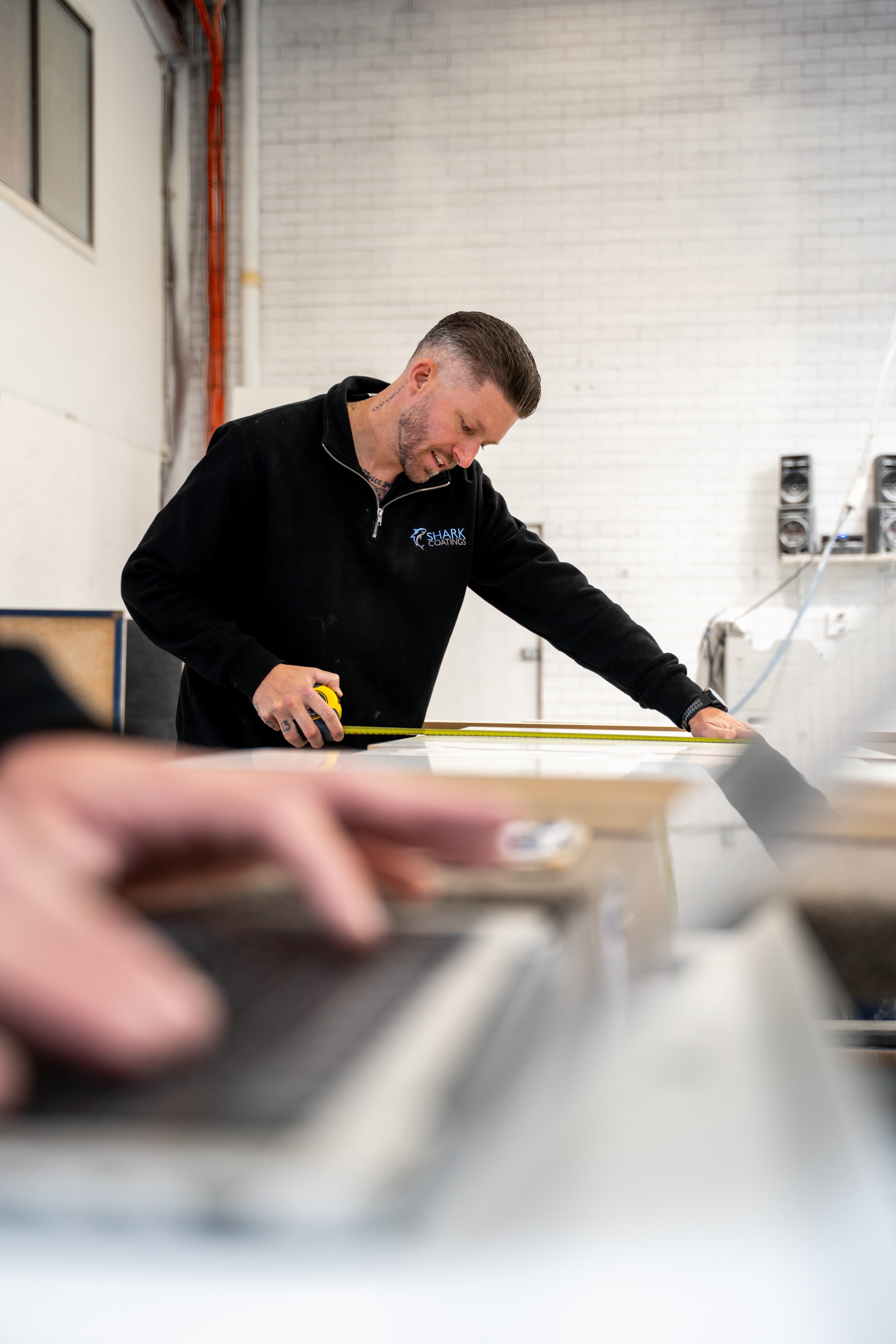 A man with tattoos on his neck, wearing a black hoodie with a shark logo, measuring a large white surface with a yellow tape measure in an industrial or workshop setting.
