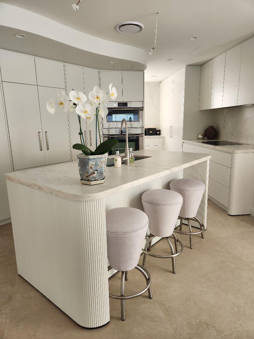 Modern white kitchen with an island, three beige barstools, a potted white orchid, built-in appliances, and minimal decor.