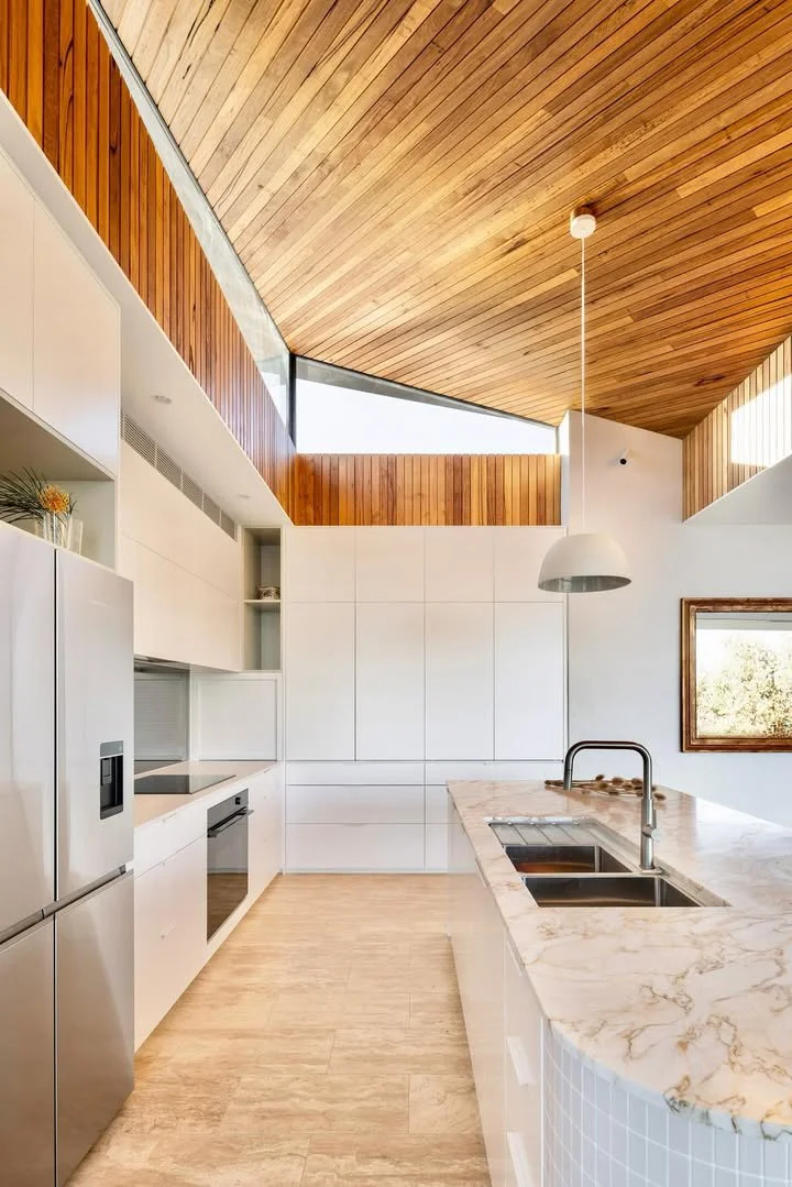 Modern kitchen with white cabinets, marble countertop island, stainless steel refrigerator, and wooden ceiling and wall accents.