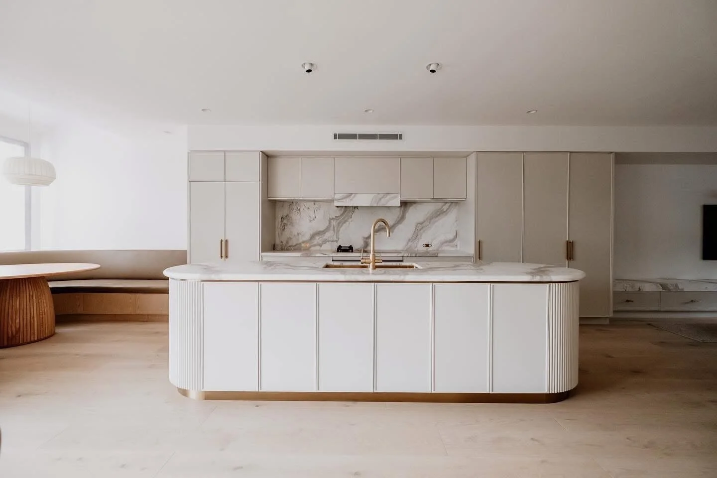 Modern, minimalist white kitchen with marble backsplash and island, featuring gold hardware and a gold faucet, surrounded by light wood flooring.