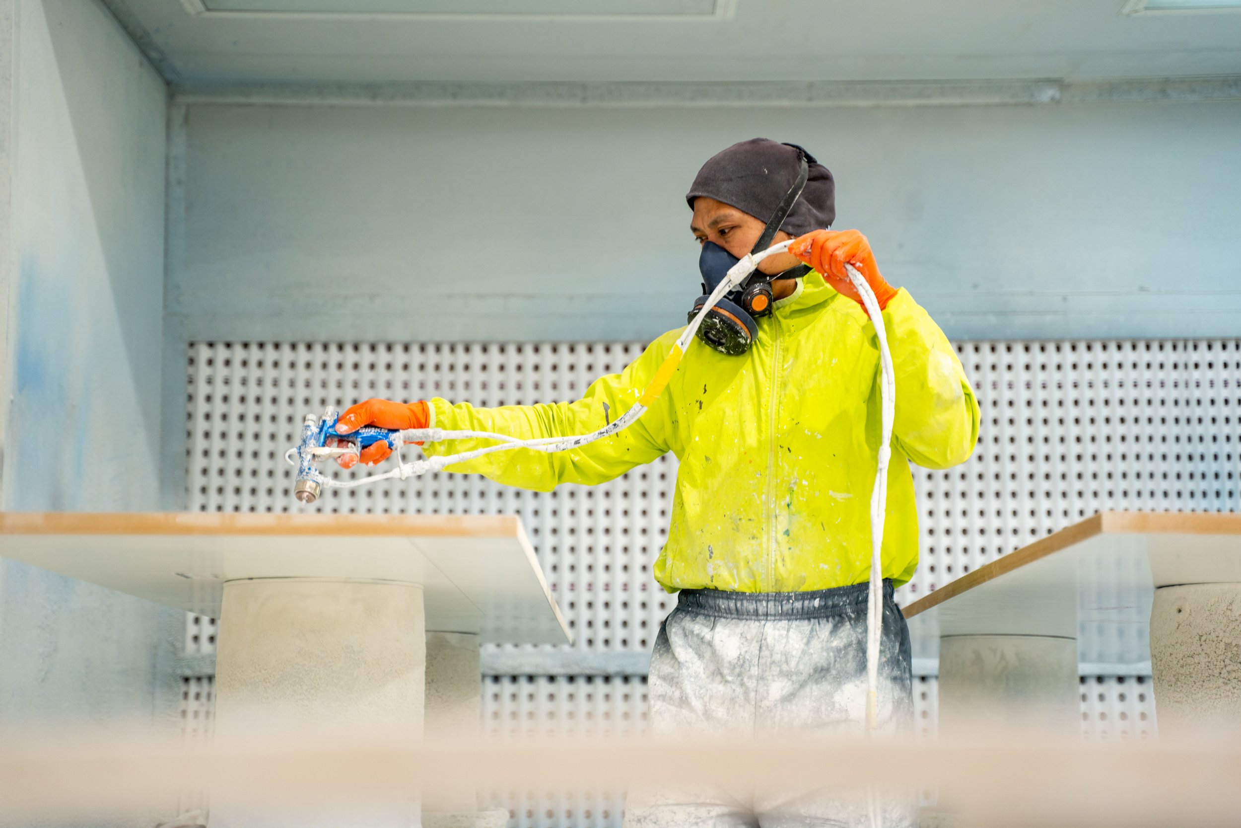 A construction worker wearing a yellow safety jacket, a dark cap, and a mask holding a spray gun, working on a light-colored piece of furniture or wall surface inside a building with a perforated wall in the background.