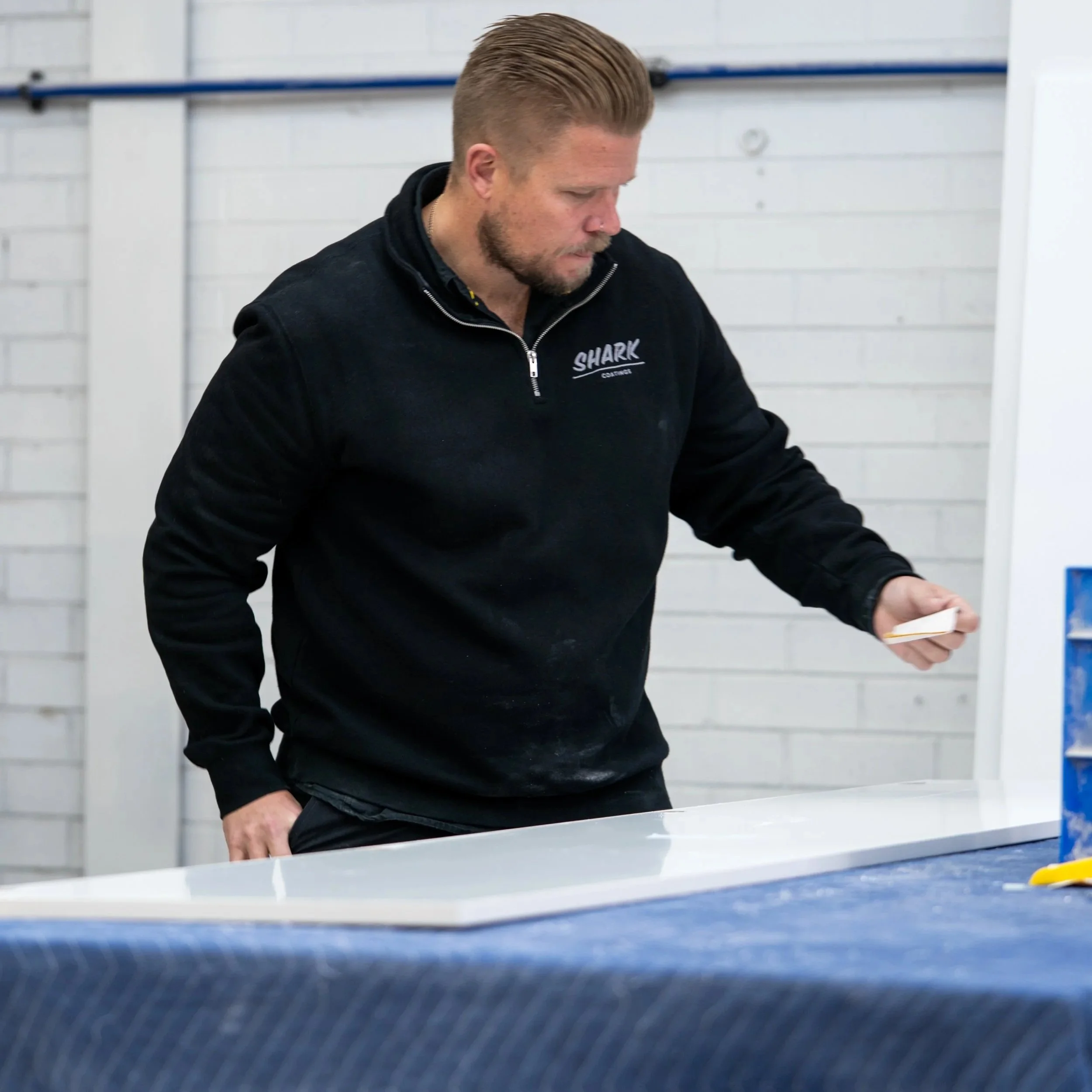 A man with styled blonde hair and a beard, wearing a black jacket with a 'SHARK' logo, is looking down and working at a table in a white-walled room, possibly an ice rink or sports facility.