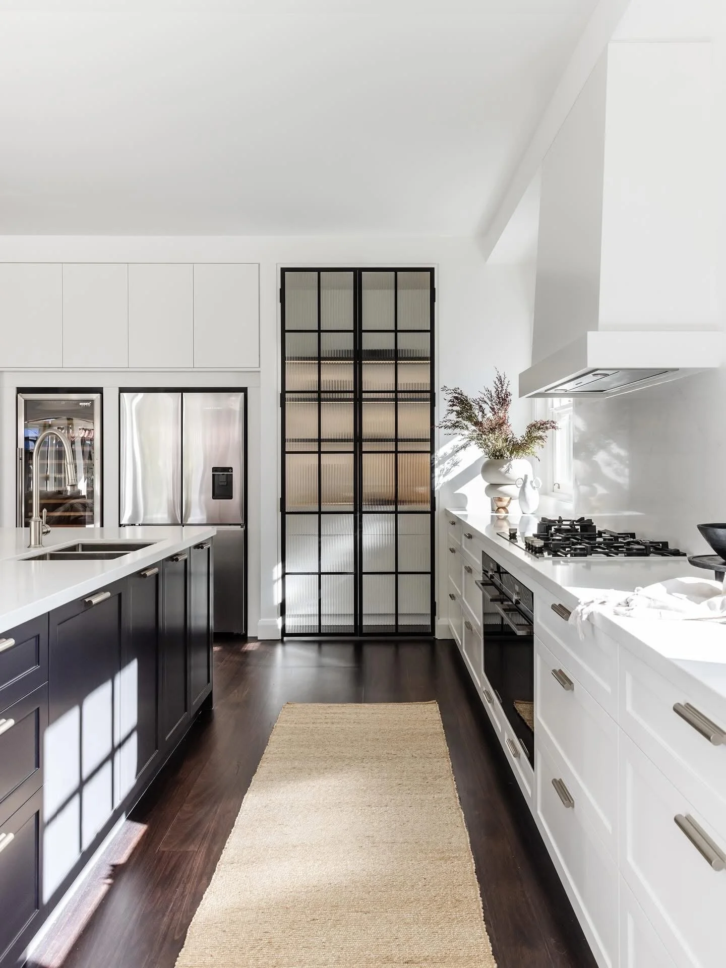 Modern kitchen with white and dark gray cabinetry, stainless steel appliances, a beige rug, a white vases with flowers, and a black-framed glass door.