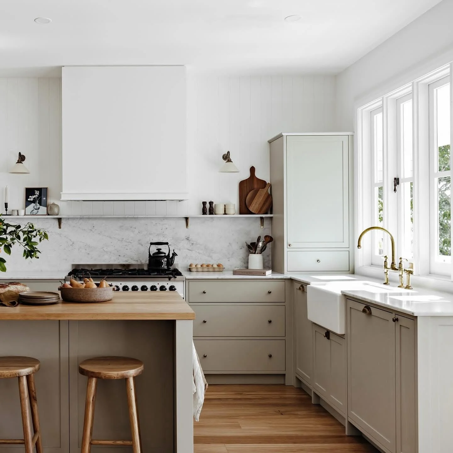 Bright white kitchen with natural wood accents, green plant, and large window with view of trees, featuring a marble backsplash, cream cabinets, and brass faucet.