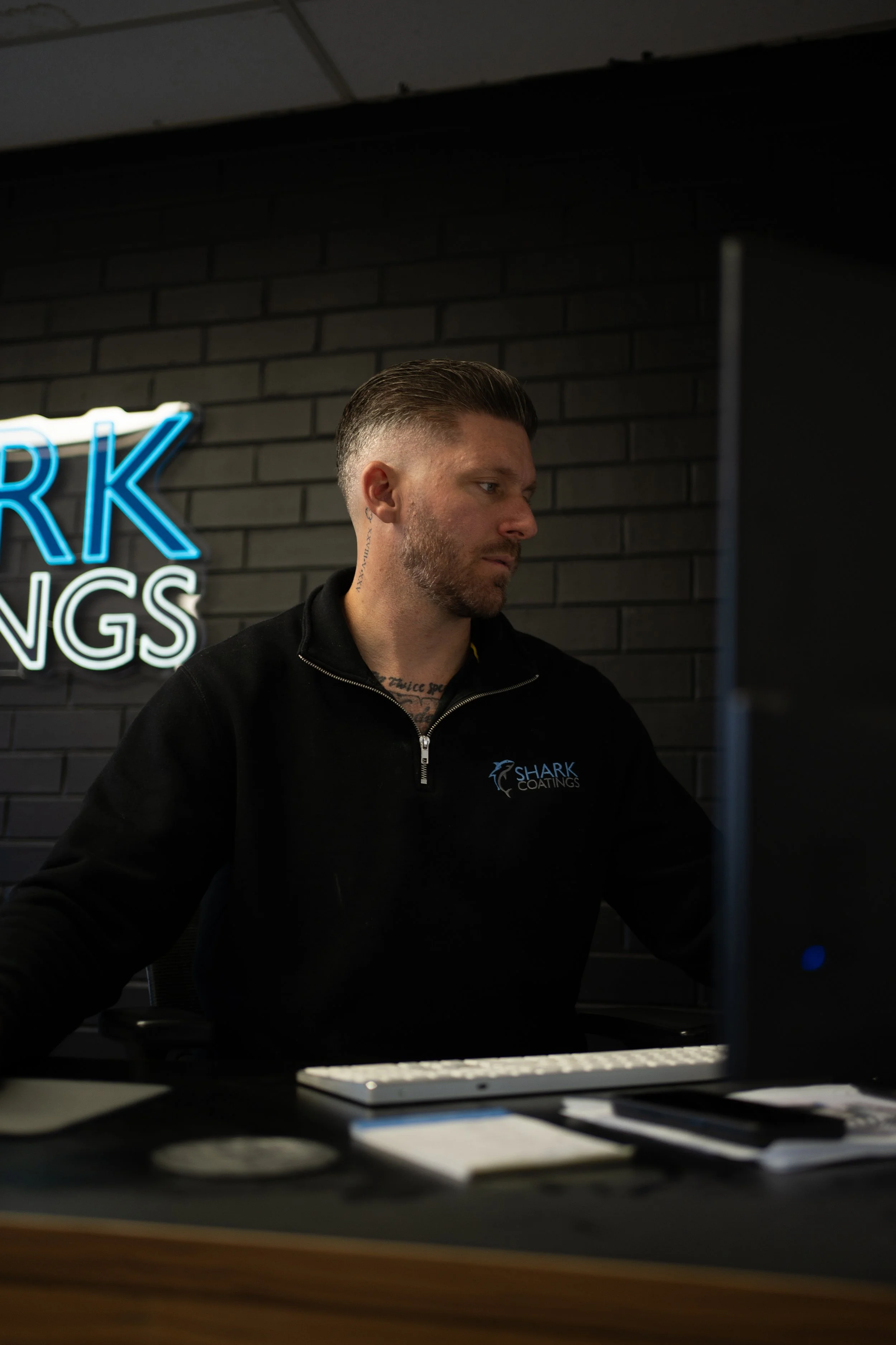 A man with short hair and a beard, wearing a black hoodie with a logo that reads 'Shark Coatings,' is sitting at a desk in front of a computer. He is in a room with dark brick walls and a neon sign partially visible that says 'ARK' and 'INGS' in blue and white. The image appears to be taken in a dimly lit office or workspace.