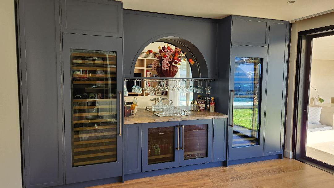A built-in blue bar cabinet with glass doors, a wine cooler, and a decorative flower arrangement on top, next to a sliding glass door.