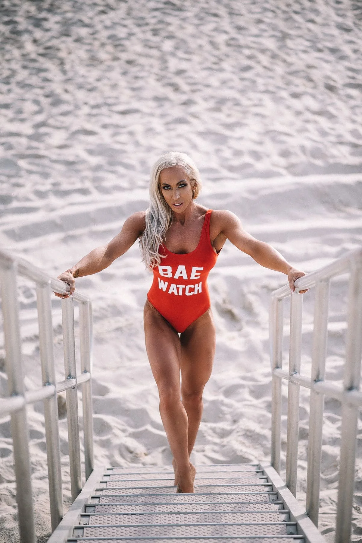  A confident model in a vibrant swimsuit gracefully walks down the stairs onto the sandy beach, showcasing her strength and fitness. 