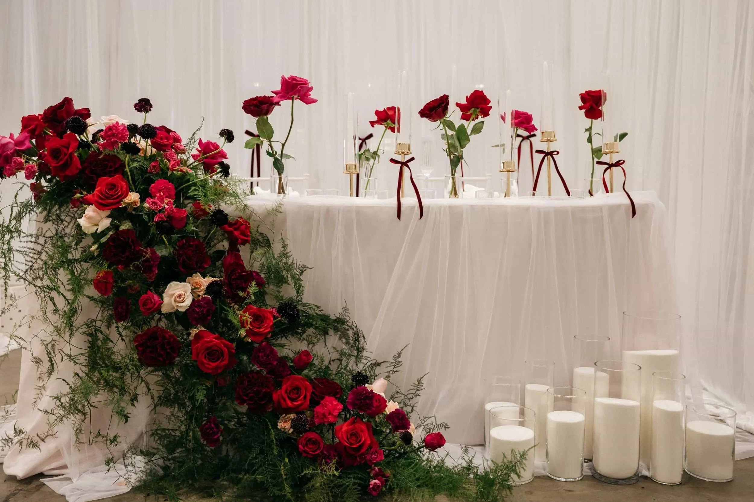 A wedding sweetheart table with a white draped backdrop, a cascading floral arrangement of red, pink, and white roses with greenery, and candles on the floor.