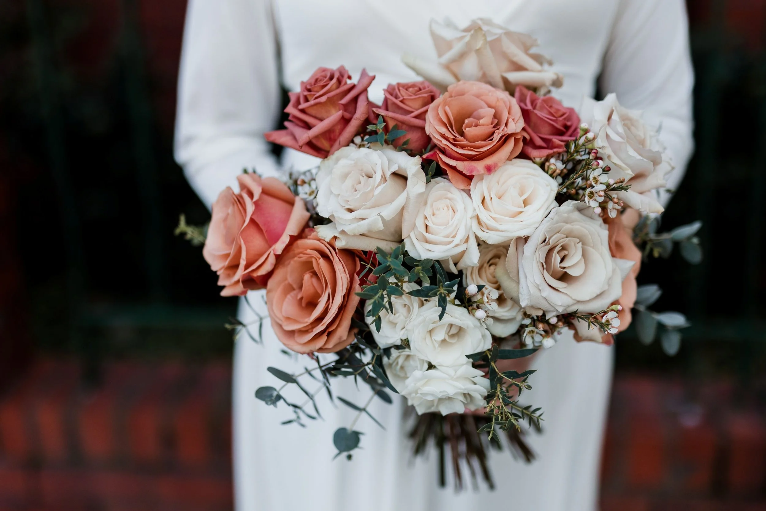 Person holding bridal bouquet with peach, white, and pink roses, with greenery and small white flowers, against a blurred dark background.