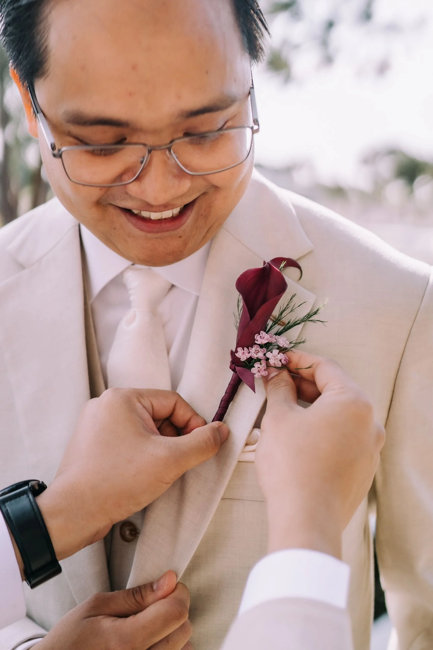 A man in a light-colored suit and glasses is smiling as someone pins a dark red boutonniere with small pink flowers and greenery onto his lapel.