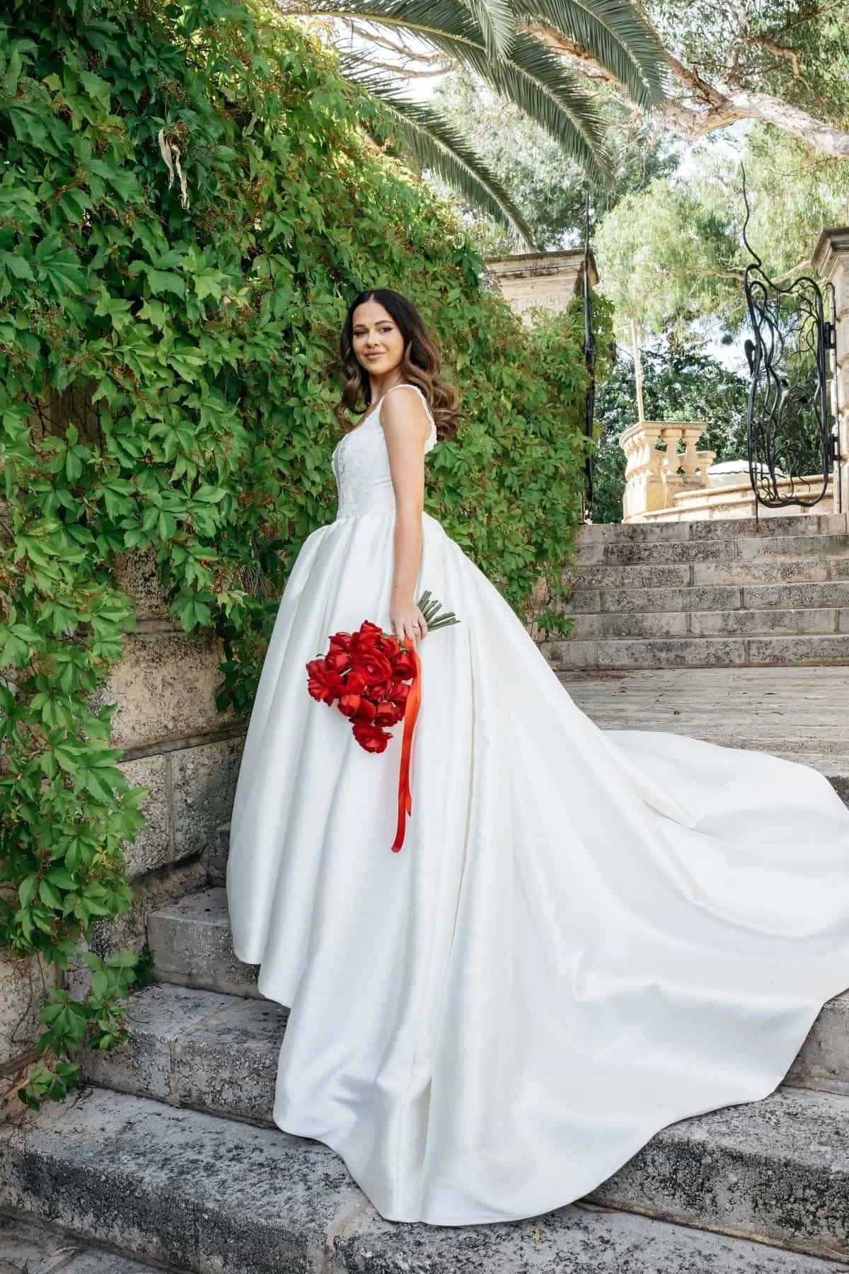 A bride in a white wedding dress holding a bouquet of red roses, standing on stone steps outdoors next to greenery and trees.