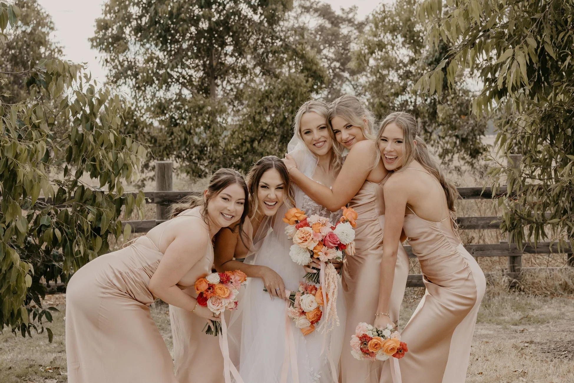 A bride and five bridesmaids celebrating outdoors, all smiling and hugging, with the bridesmaids holding bouquets of orange, pink, and white flowers, surrounded by trees and a wooden fence.