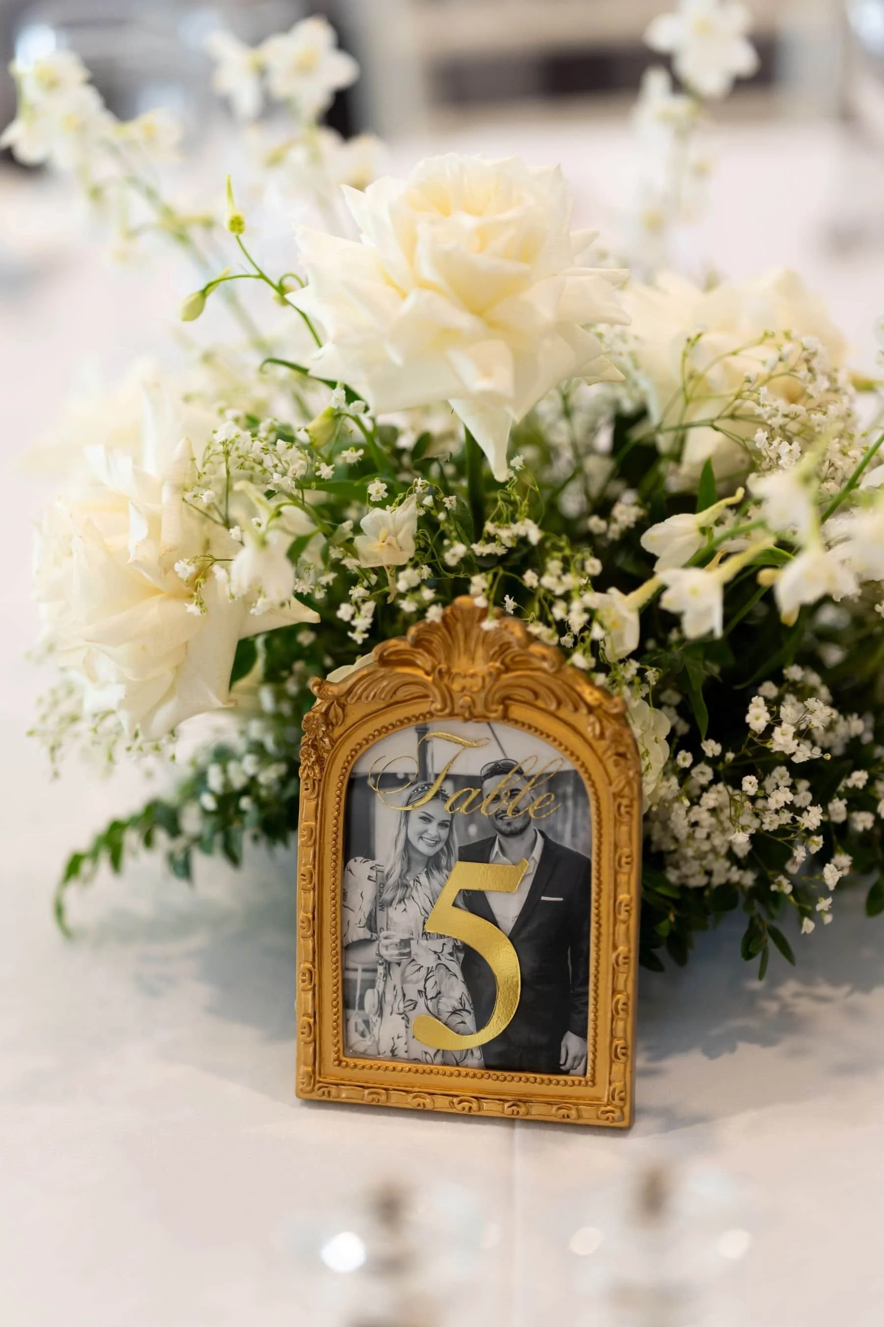 A floral centrepiece with white roses and baby's breath, featuring a framed photo with a gold number 5 in front of it, on a white table.