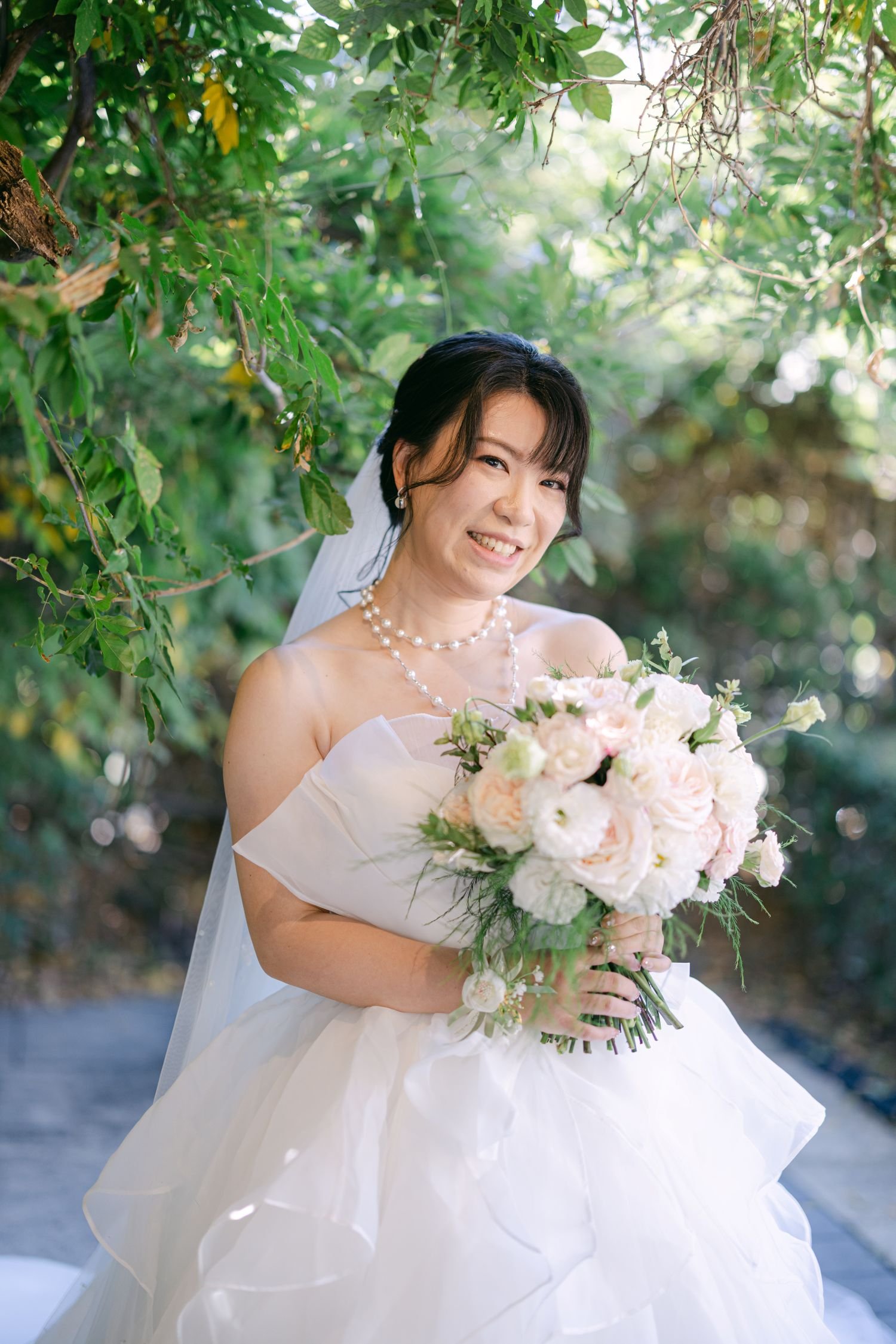 A smiling bride with dark hair styled in an updo, wearing a strapless white wedding dress, pearl necklace, and earrings, holding a large bouquet of white and blush pink flowers, standing outdoors among green trees and foliage.