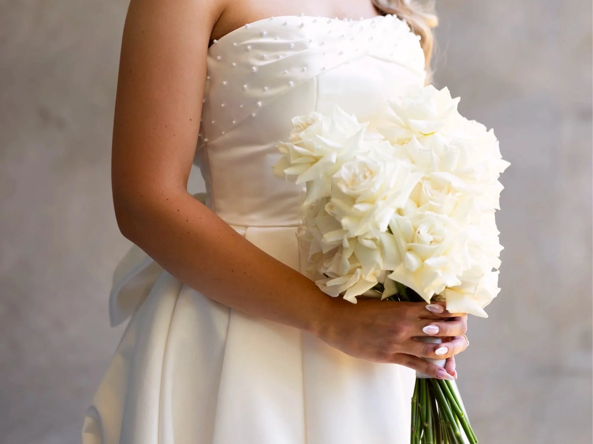 A bride in a white wedding gown holding a large bouquet of white roses and calla lilies, against a neutral background.