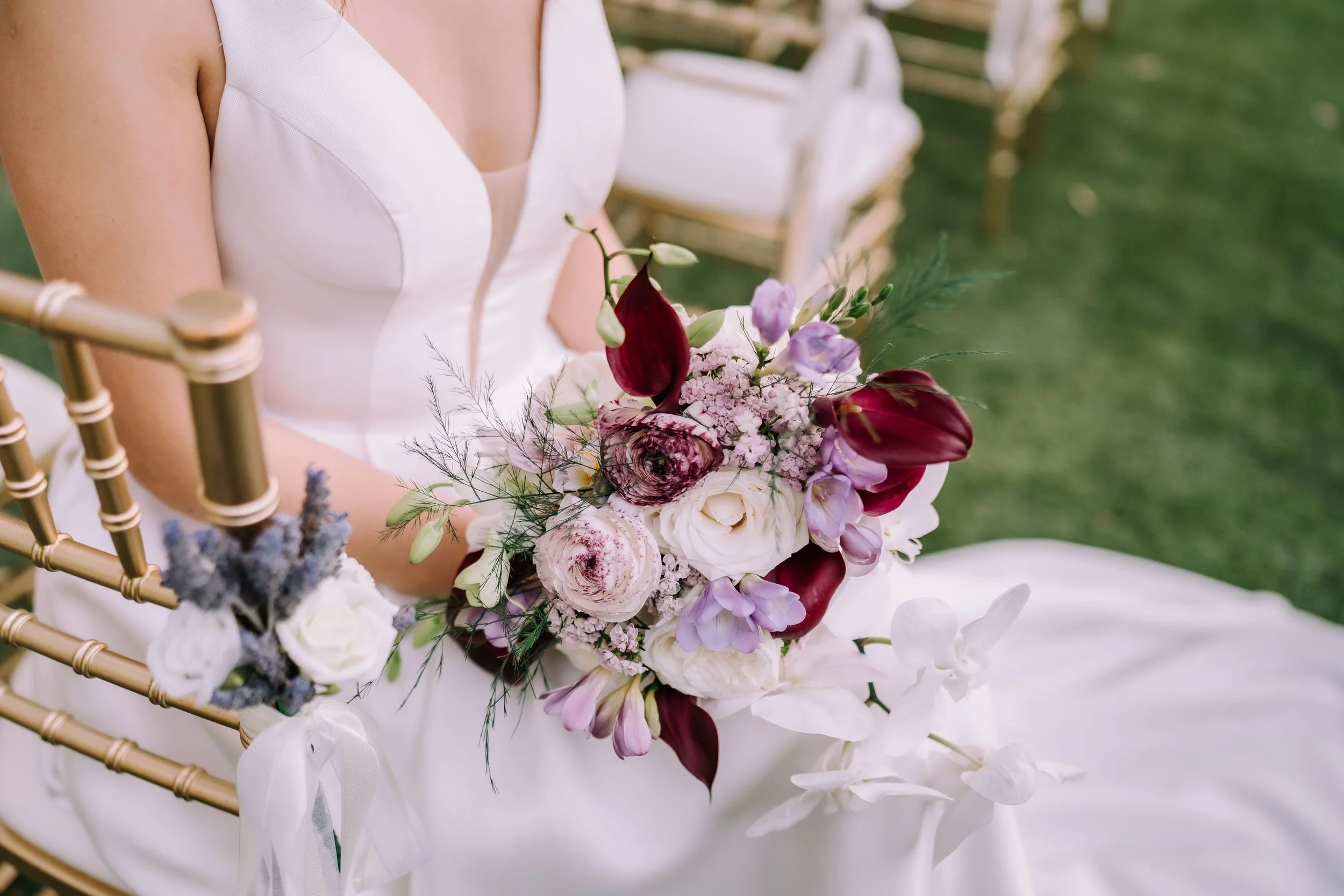 Close-up of a wedding bouquet with white, pink, purple, and deep red flowers, held by a woman in a white dress, seated on a gold chair outdoors.