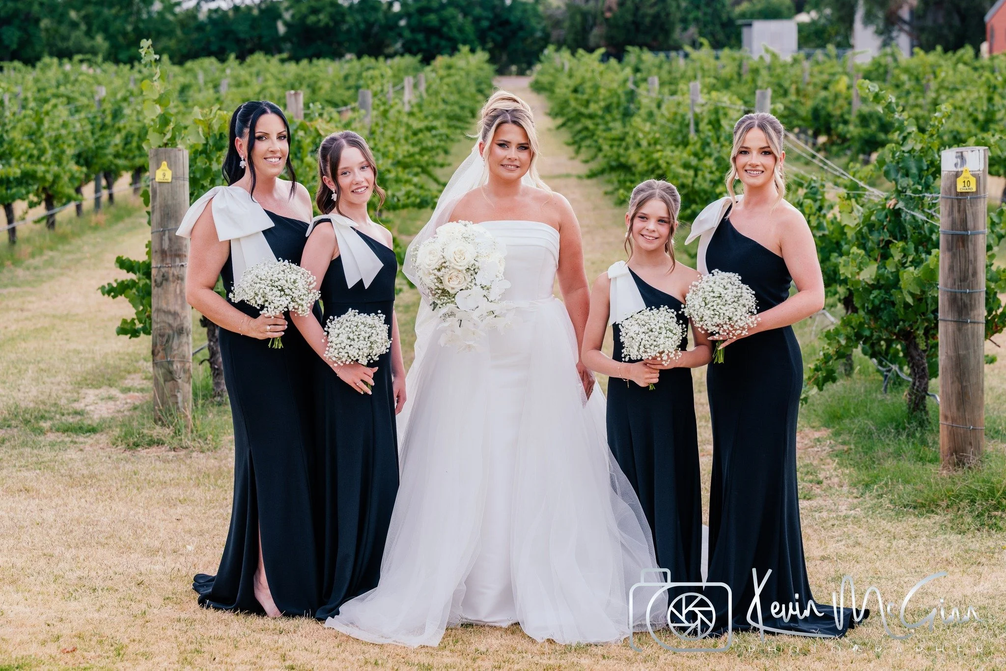 A group of five women, including a bride in a white wedding dress holding a bouquet, and four bridesmaids in matching black dresses with white bows, standing in a vineyard.