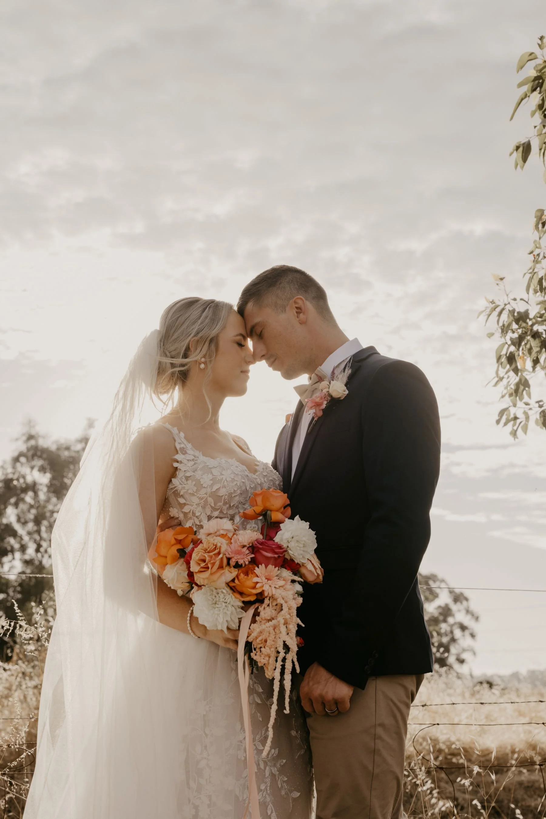 A bride and groom standing close together outdoors, touching foreheads, with a cloudy sky and trees in the background. The bride wears a white lace wedding dress and holds a colorful bouquet, while the groom wears a black suit, white shirt, and a bou