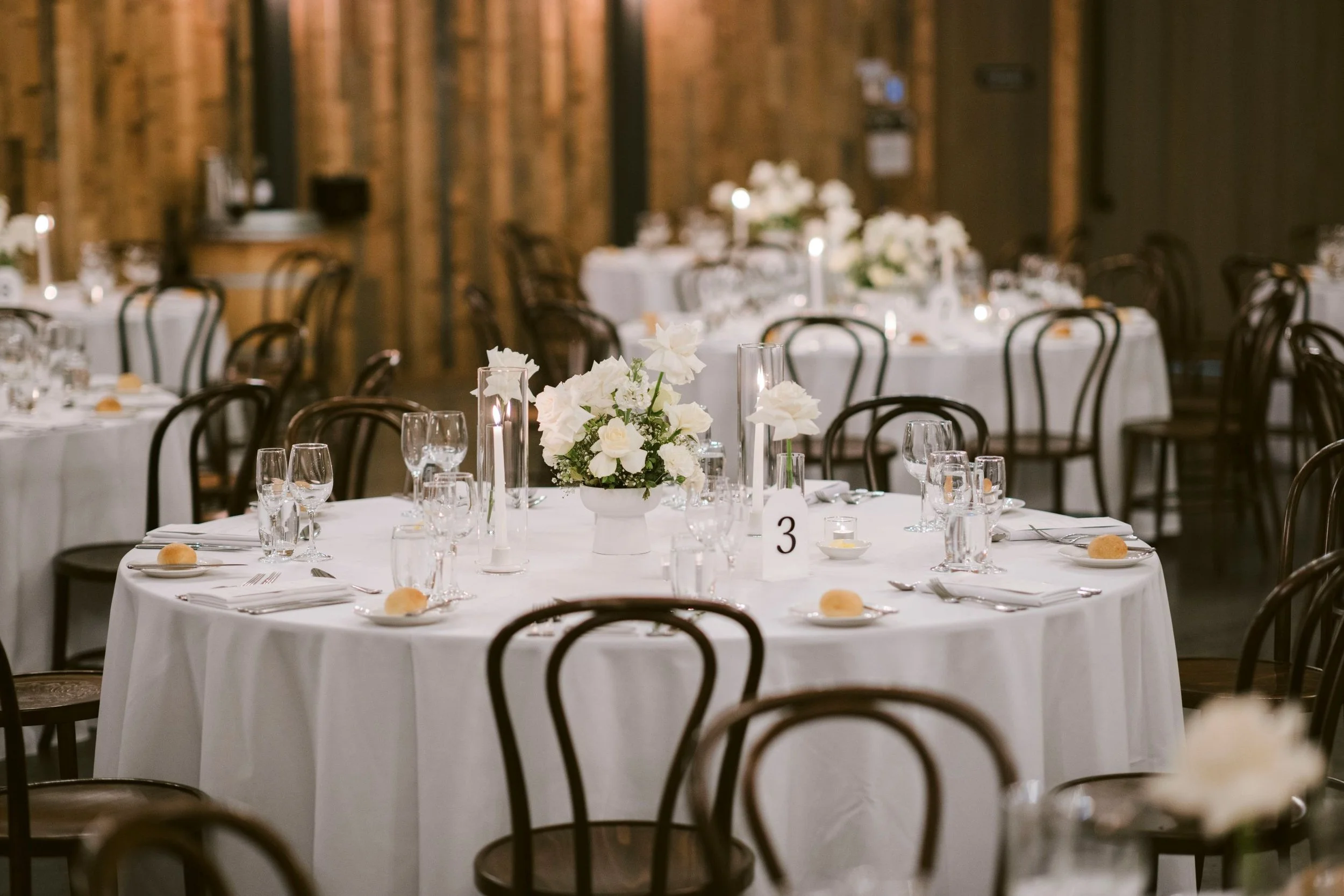Elegant banquet hall with round tables covered in white tablecloths, decorated with white floral centerpieces and lit candles, set for a formal event.