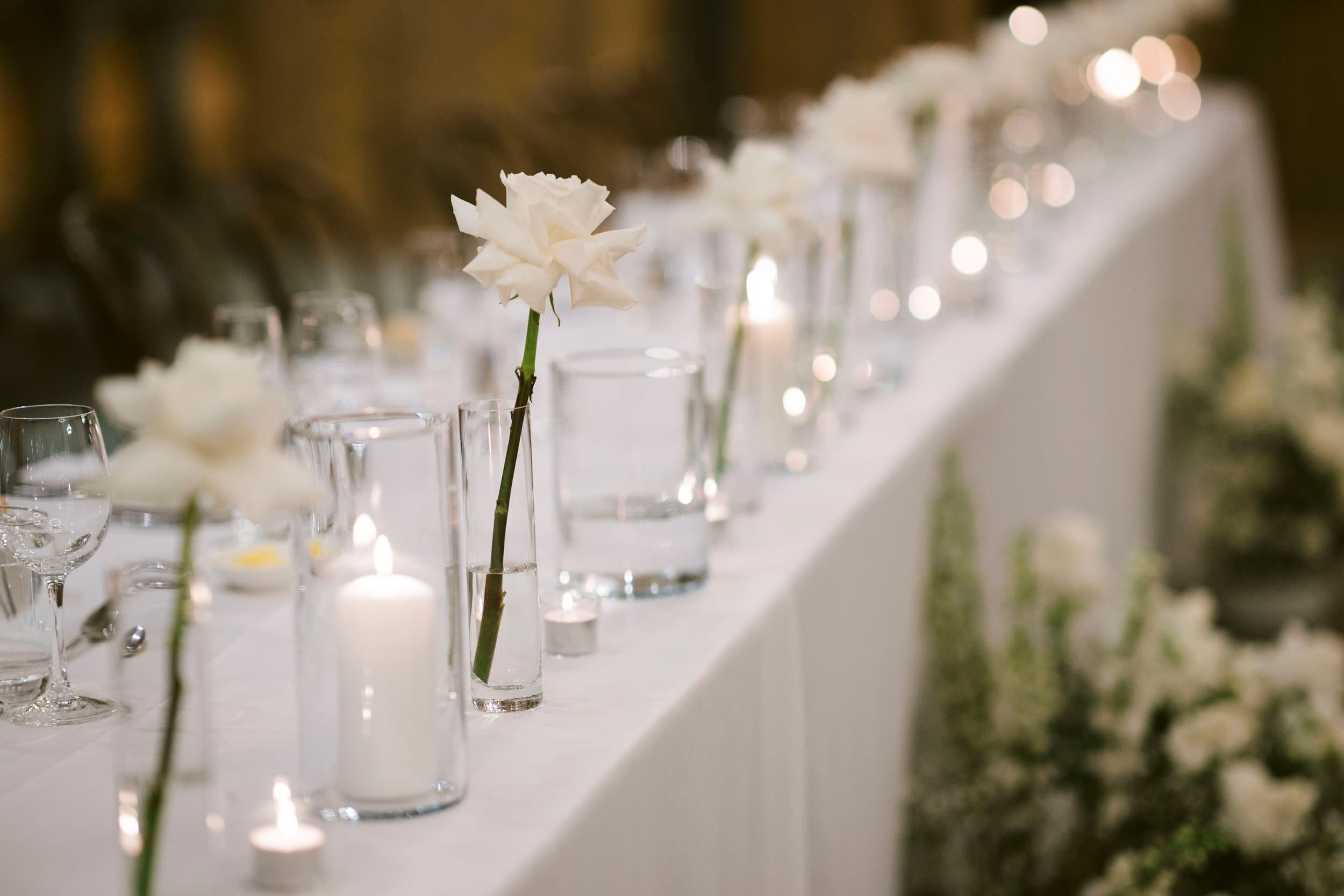 White rose bud vases on a bridal table set for an wedding, with bottles and candles in the background.