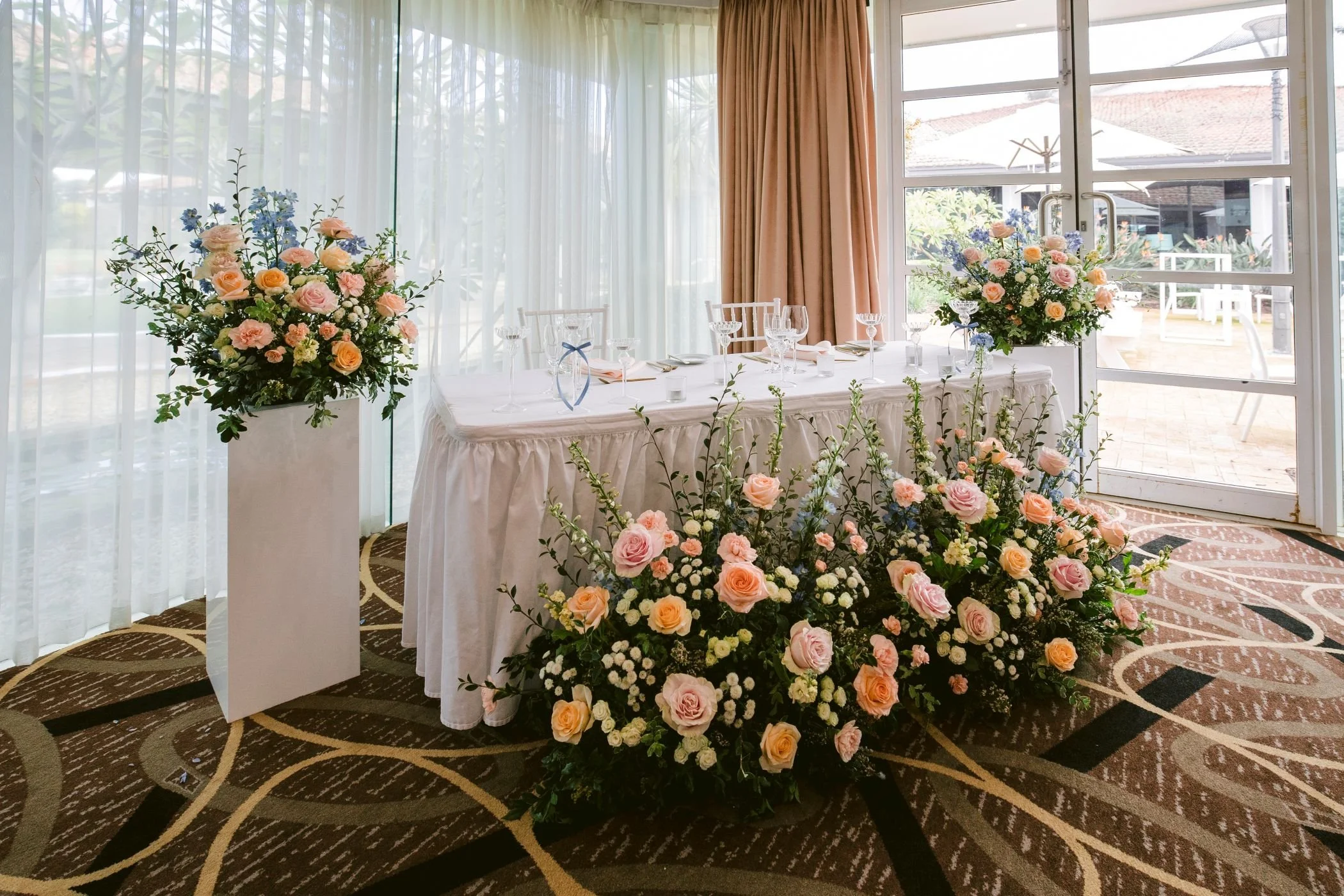 Wedding or event table setup with large flower arrangements of pink, peach, and blue flowers, with glassware and table settings, in a room with large windows and curtains.
