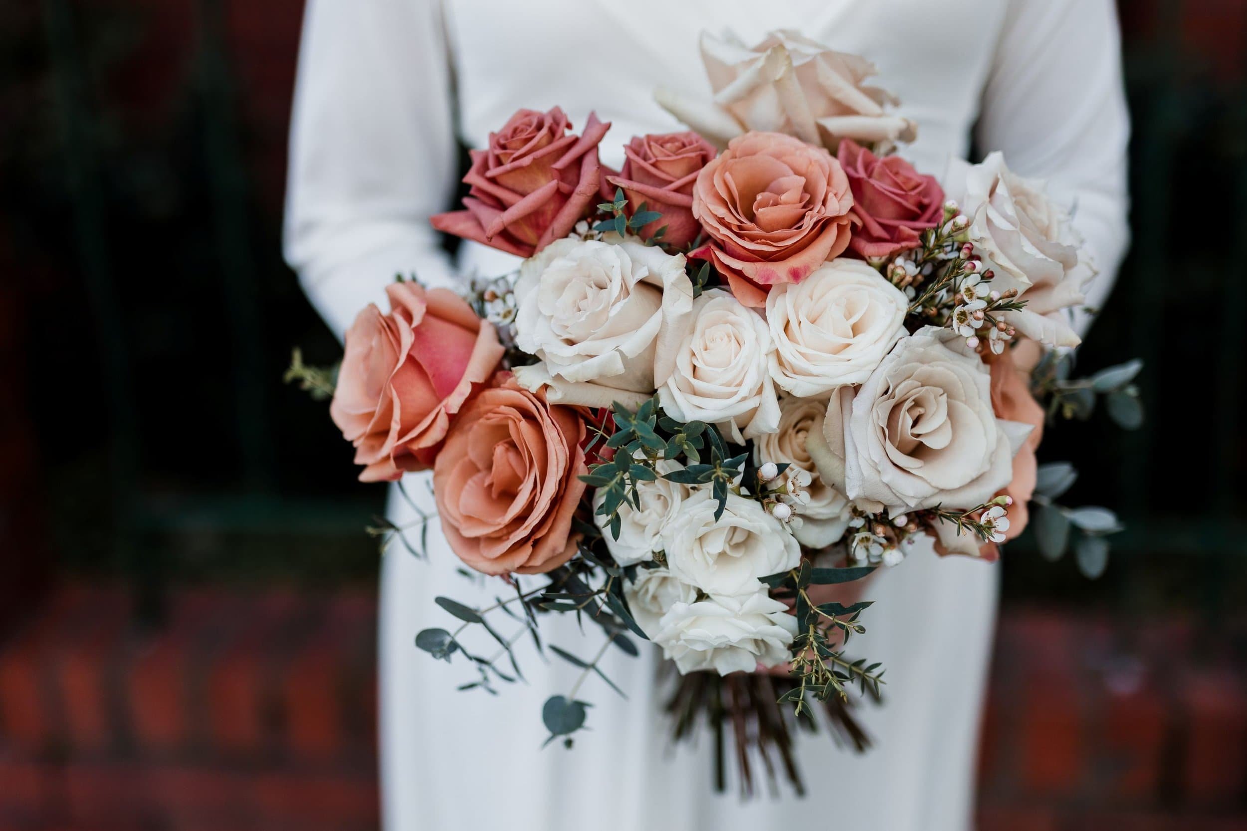 A bride holding a bouquet of pink, cream, and peach roses with greenery, wearing a white shirt or dress, against a blurred dark background.